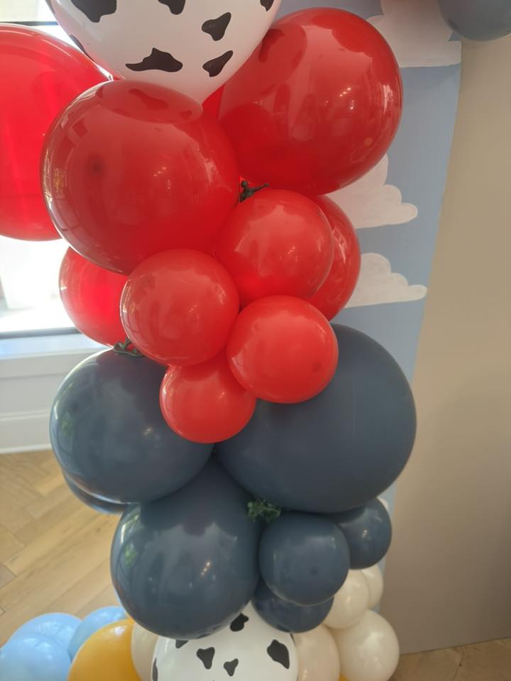 Toy Story themed balloon garland with red, navy blue, and white cow-print balloons against a cloud backdrop.