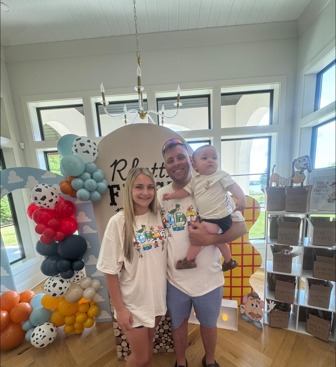 Family wearing Toy Story themed t-shirts standing in front of a party display with balloon arches and gift boxes.