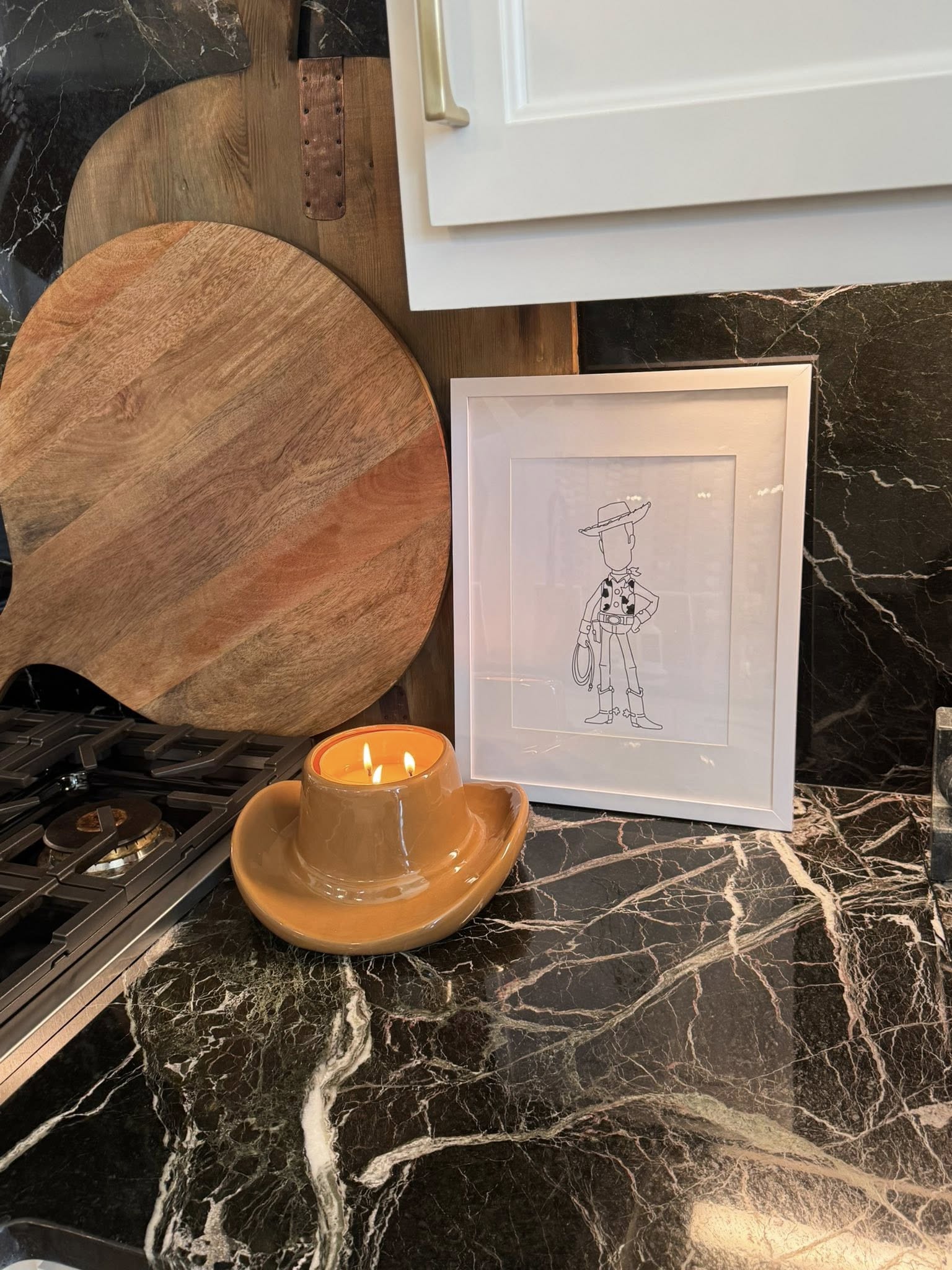 Cowboy hat shaped candle lit on a black marble countertop next to a framed Woody line drawing.