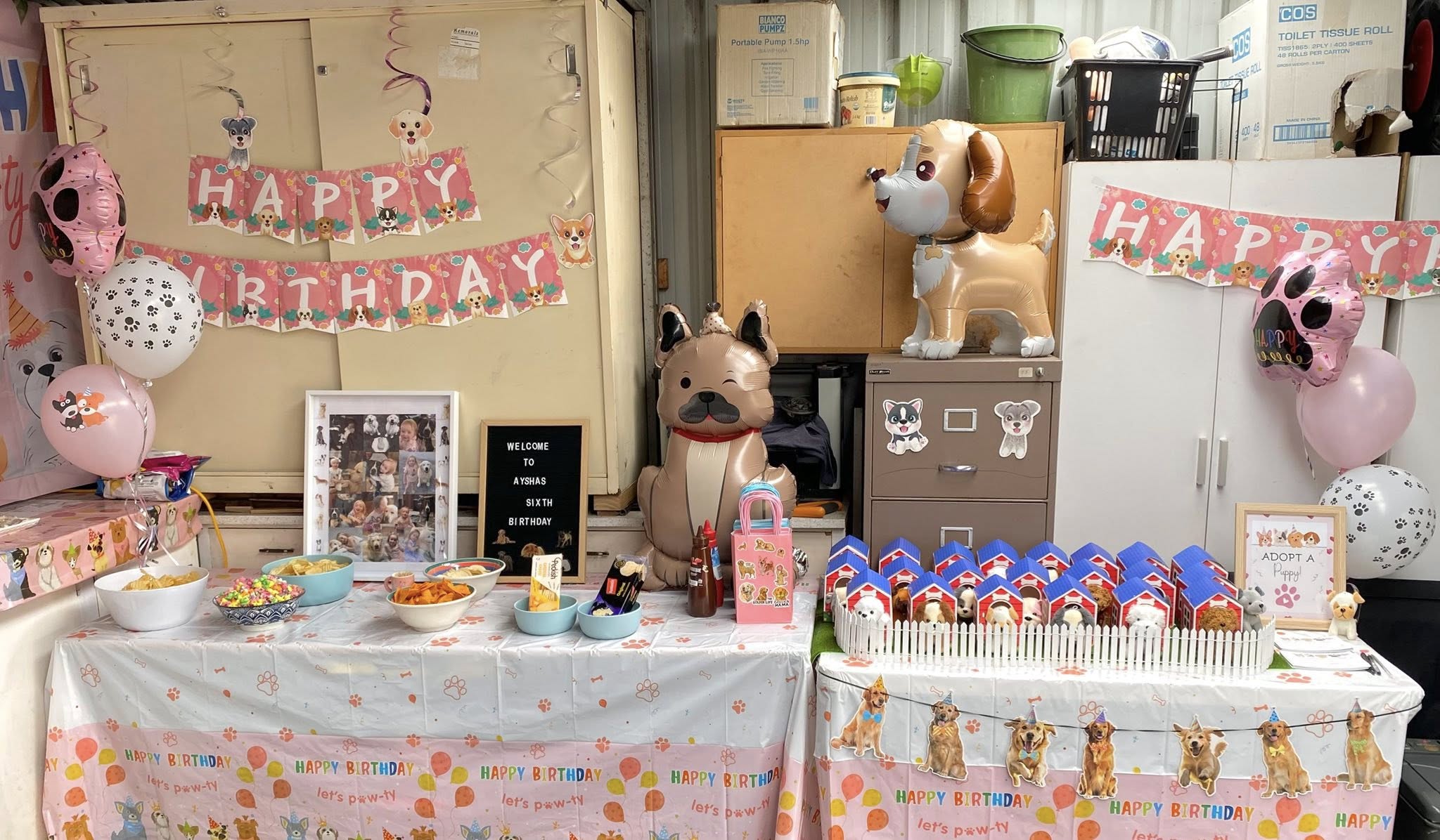 Puppy-themed birthday party decorations with balloons, photo frame, snacks, and toy dog houses on tables.