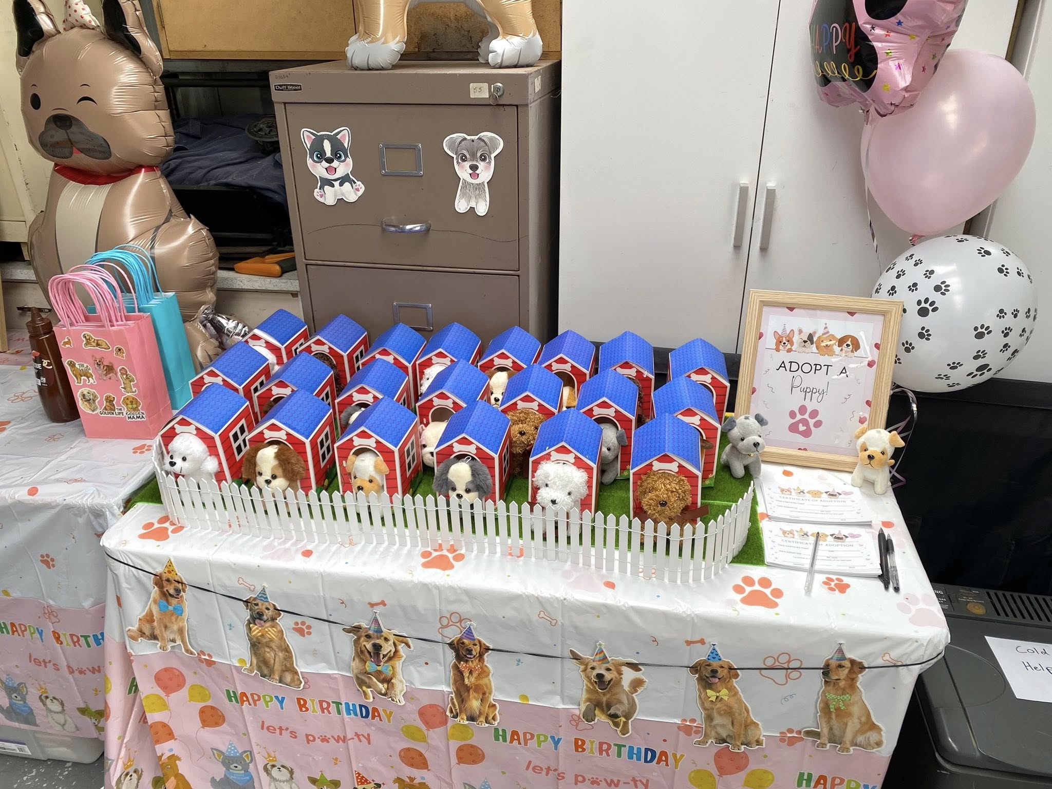 Puppy adoption birthday party display featuring plush dogs in small red kennel boxes behind a white picket fence