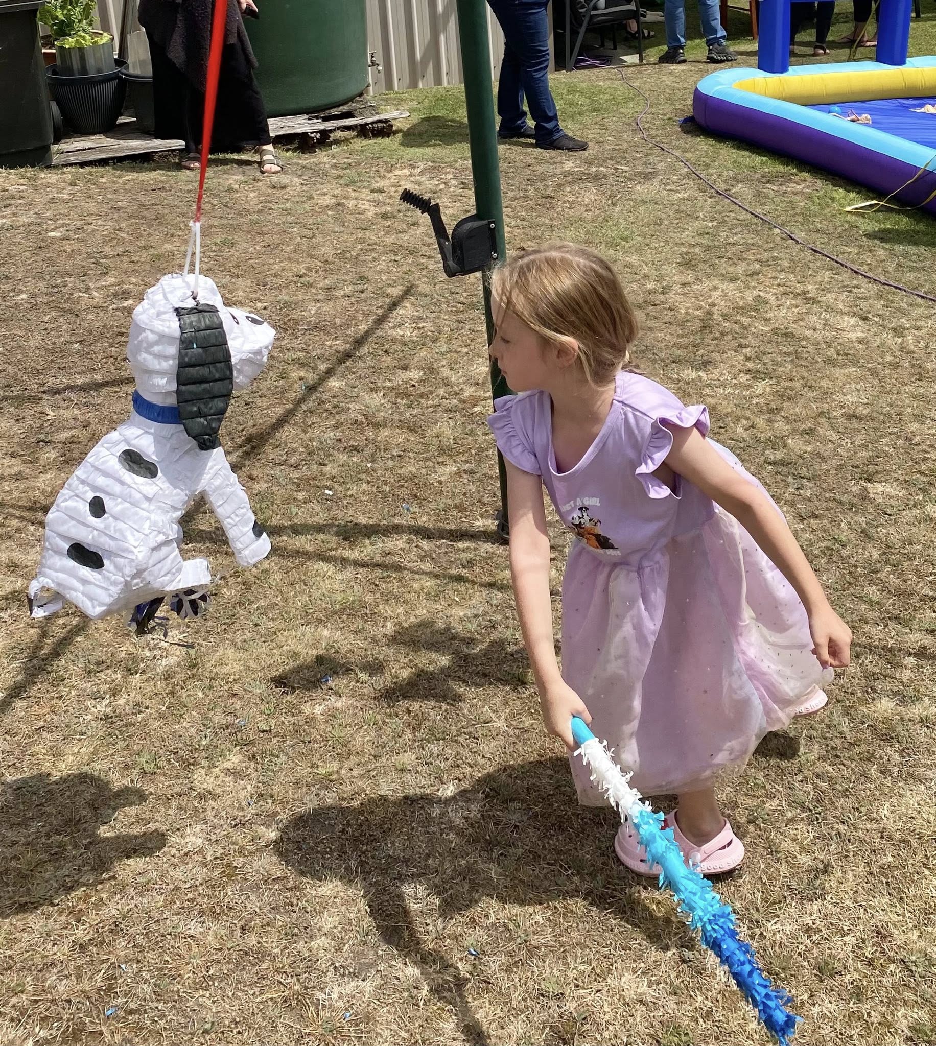 Young girl in a purple dress hitting a Dalmatian-shaped dog pinata at an outdoor party