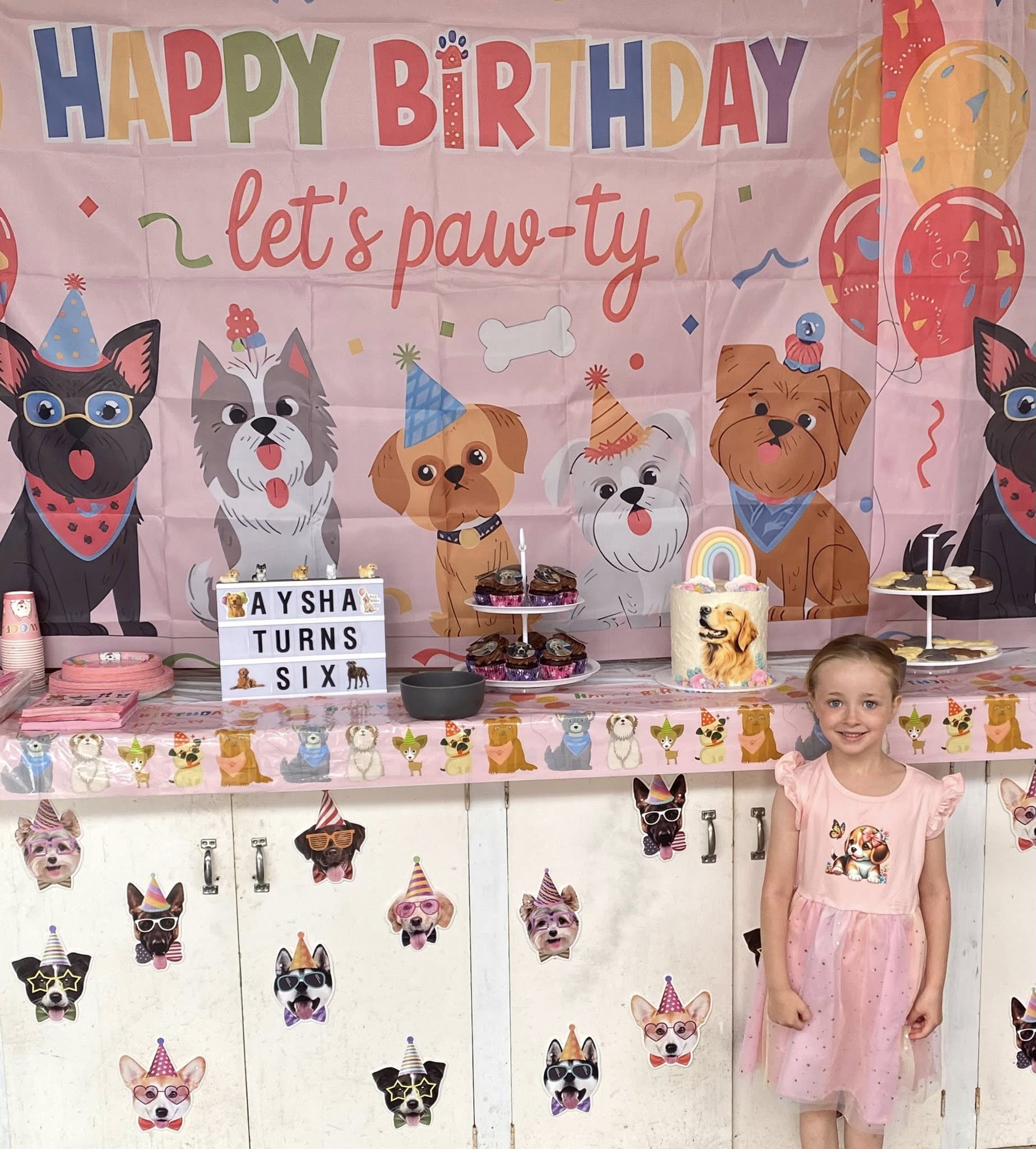 Young girl smiling by a pink dog-themed birthday party table with decorations and a rainbow cake
