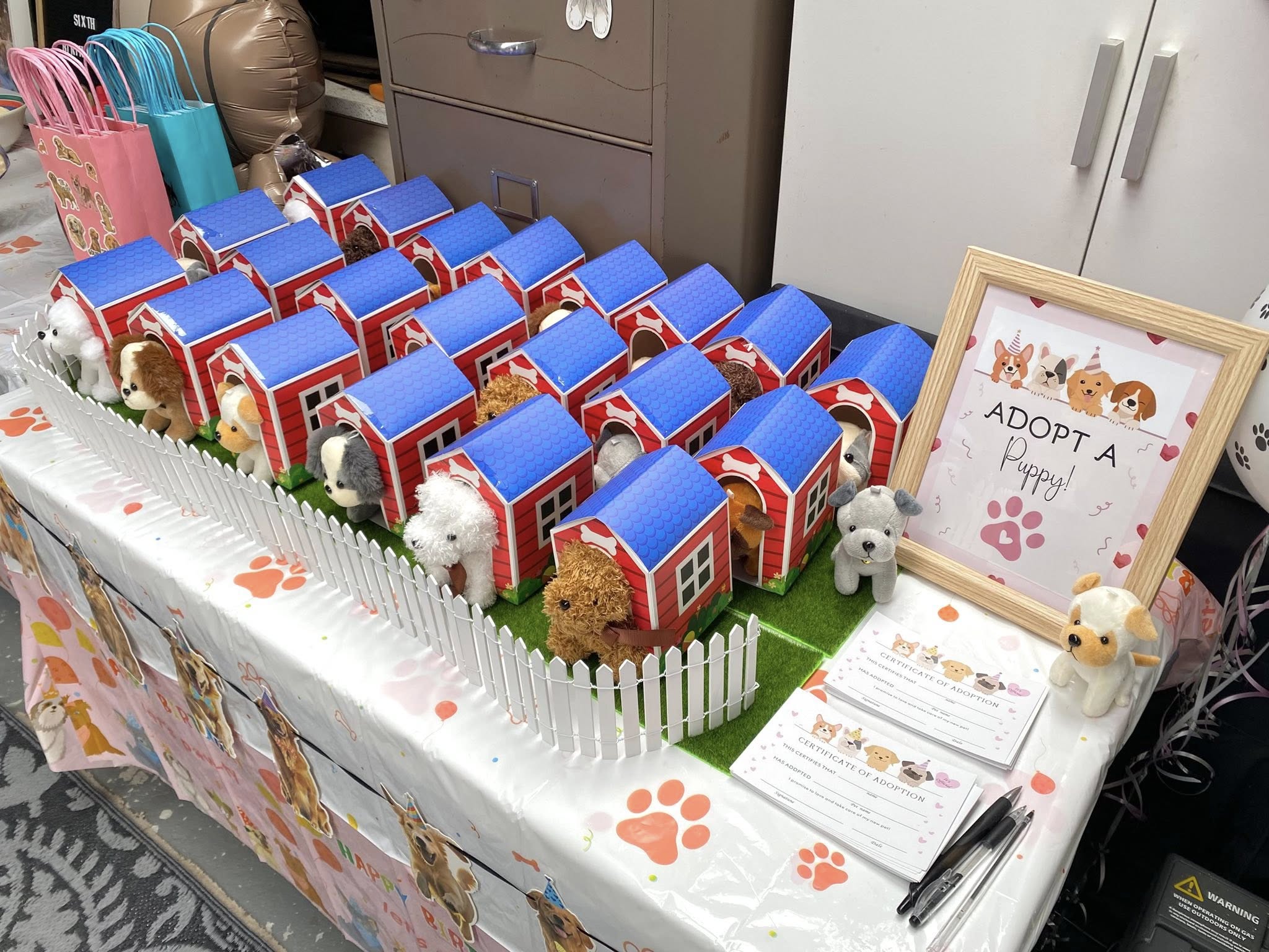 Adopt a Puppy party favor display featuring plush puppies in individual red cardboard dog houses with white picket fencing and adoption certificates.