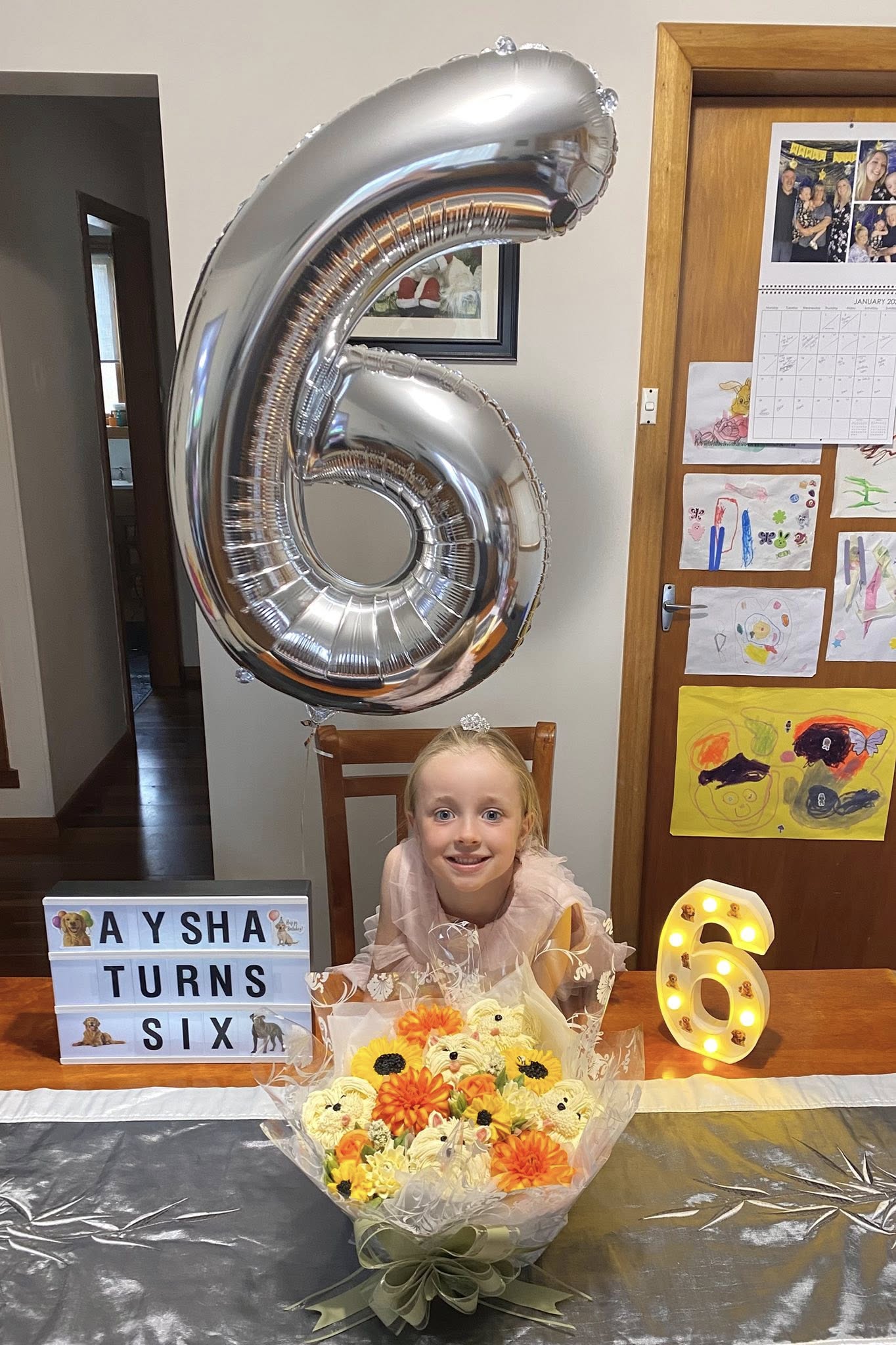 Young girl celebrating her sixth birthday with a large silver number balloon, light-up sign, and cupcake bouquet.