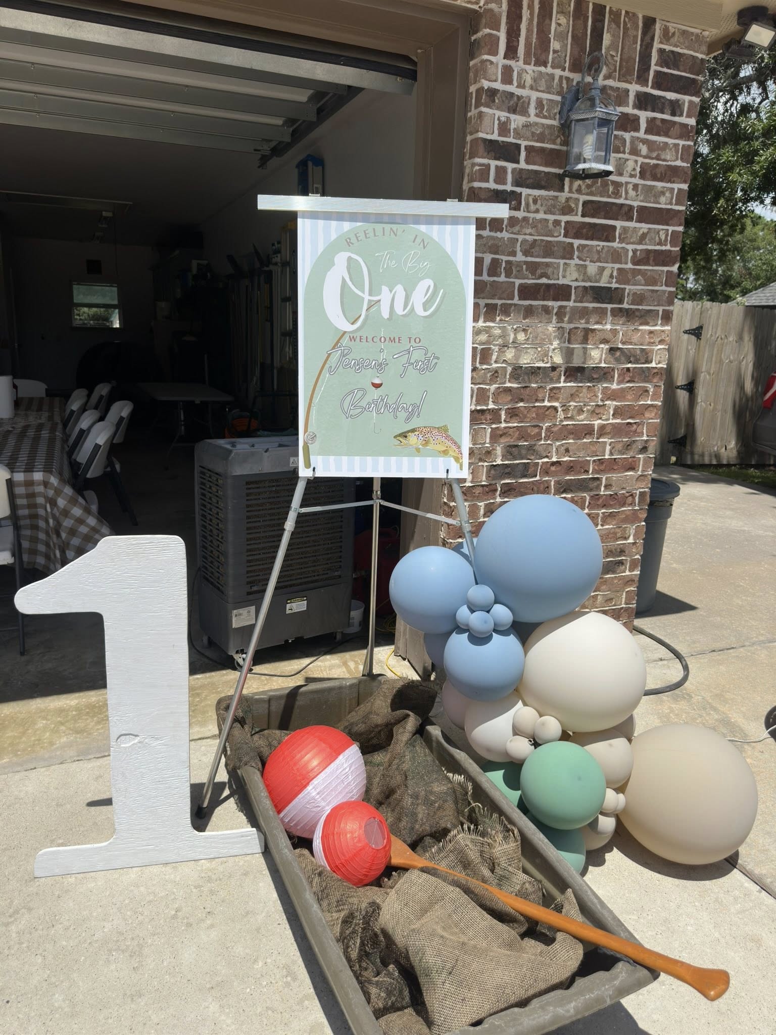 Reelin' In The Big One first birthday party sign with blue balloons and a fishing-themed wagon display.