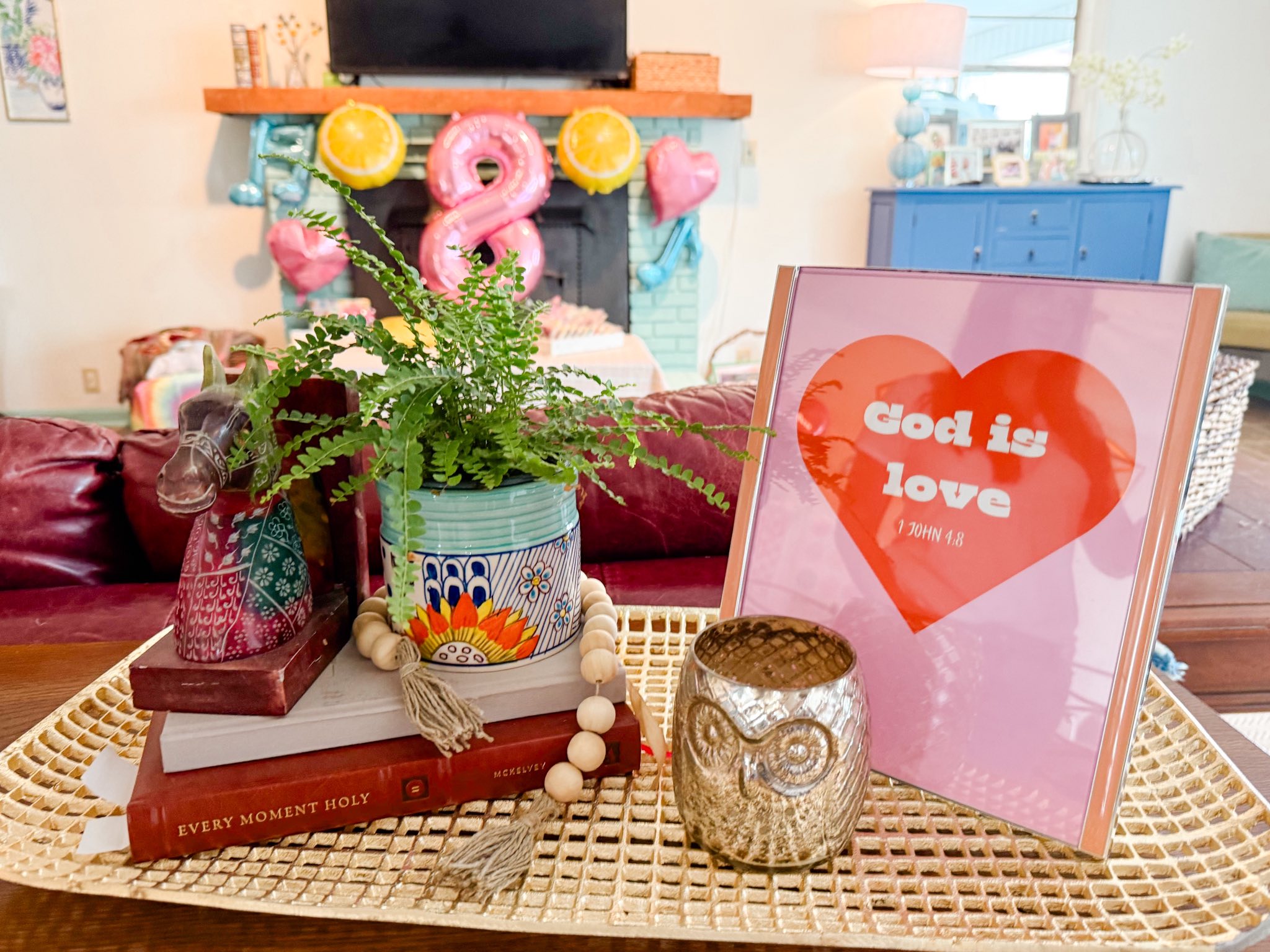 Tabletop decor featuring a God is Love sign, a fern in a patterned pot, an owl votive, and stacked books.