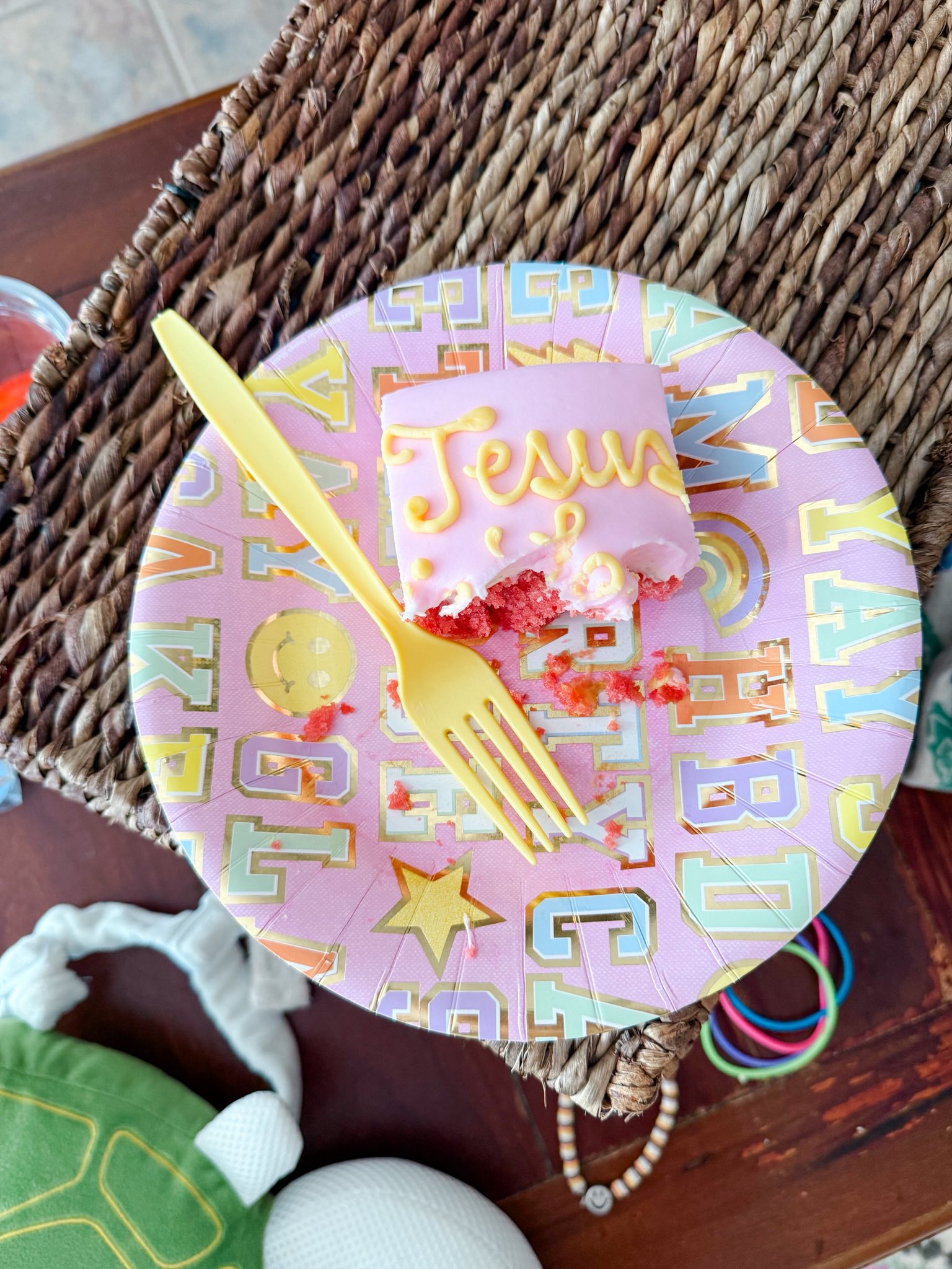 A bite-taken square cake slice with pink frosting and 'Jesus' written in yellow icing on a patterned party plate with a yellow fork.