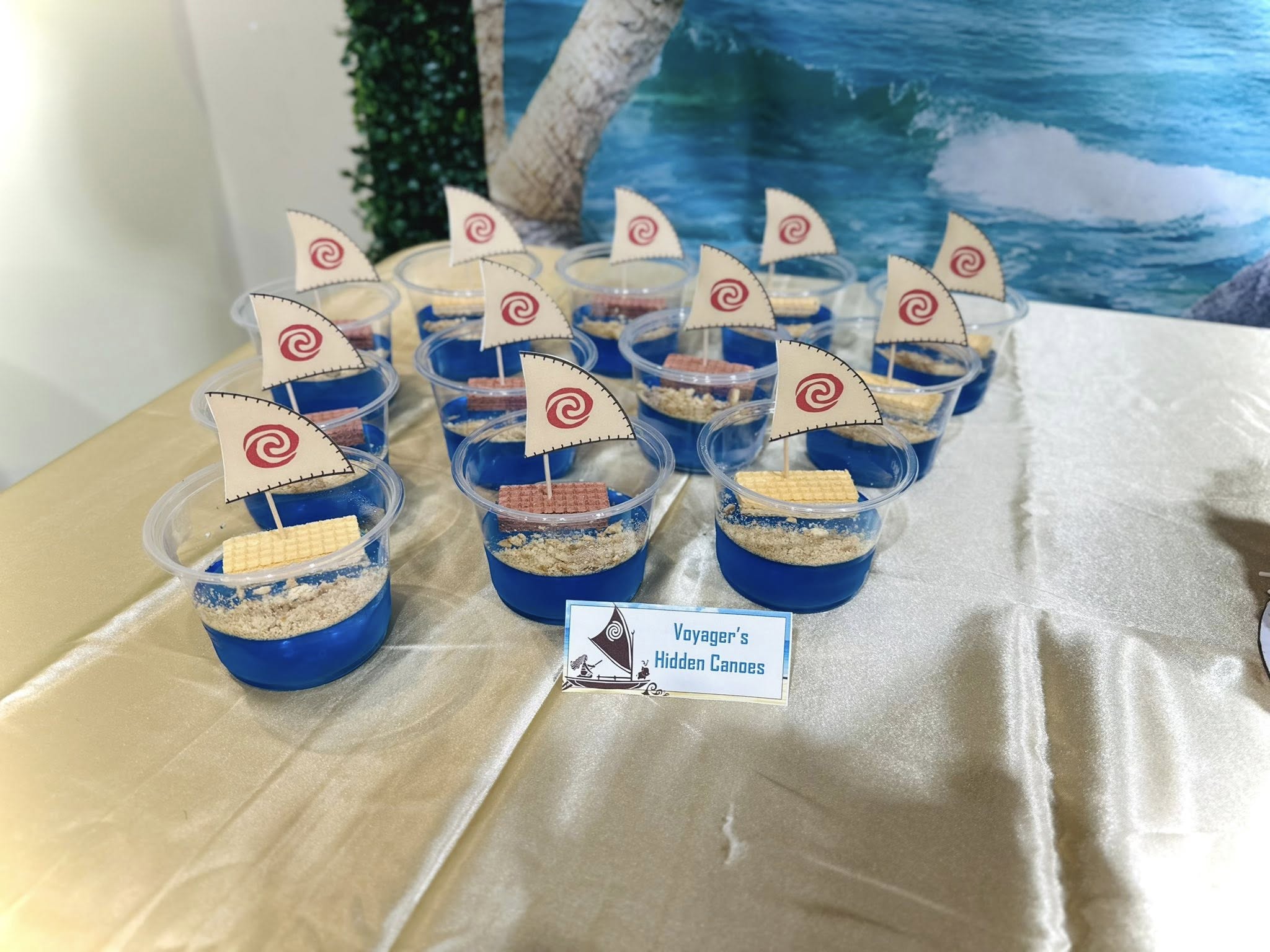 Moana-themed cups filled with blue jello and cookie crumb sand, topped with small sail decorations labeled Voyager's Hidden Canoes.