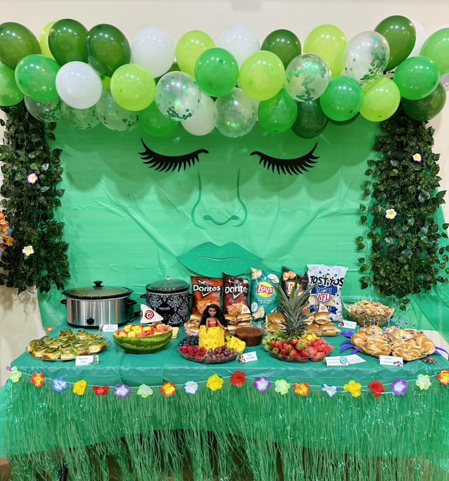 Moana themed party food table with green balloon arch, tropical decor, assorted snacks, and fruit platters.