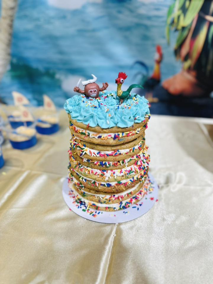 Layered cookie cake with sprinkles, topped with bright blue frosting and Moana-themed figurines.