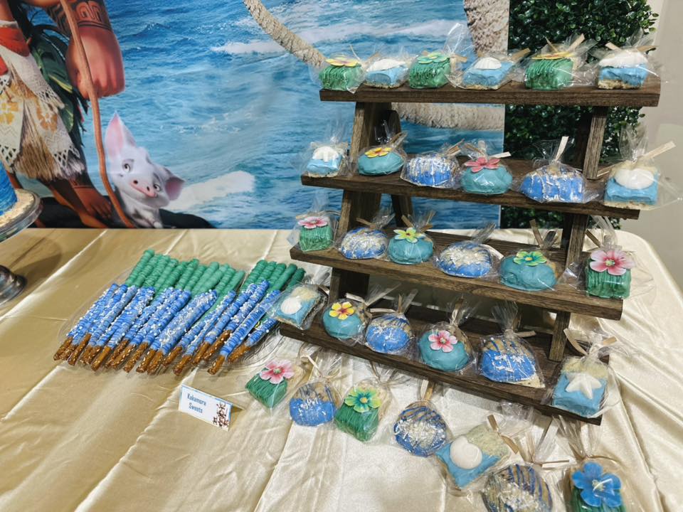Moana themed chocolate-covered pretzel rods and individual packaged tropical sweets on a tiered display stand.