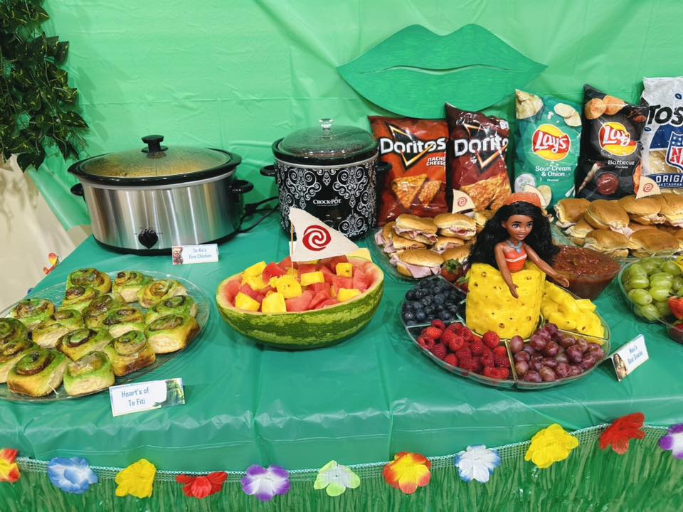 Table set with snacks and Moana-themed food including a watermelon boat and fruit platter decorated with a Moana doll.
