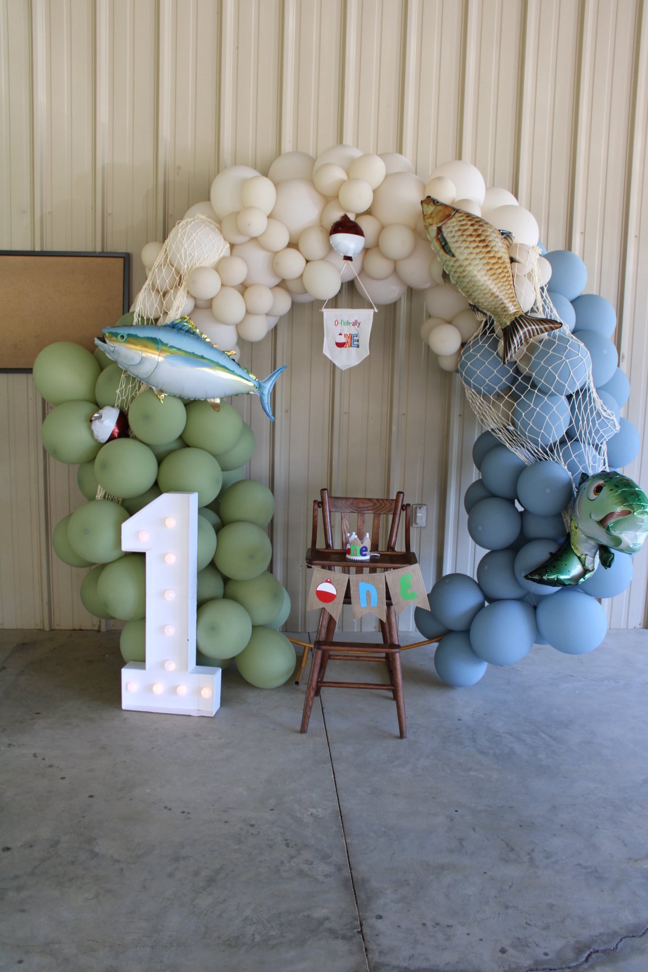 Fishing-themed first birthday party setup with a balloon arch, a light-up number one, and a high chair.