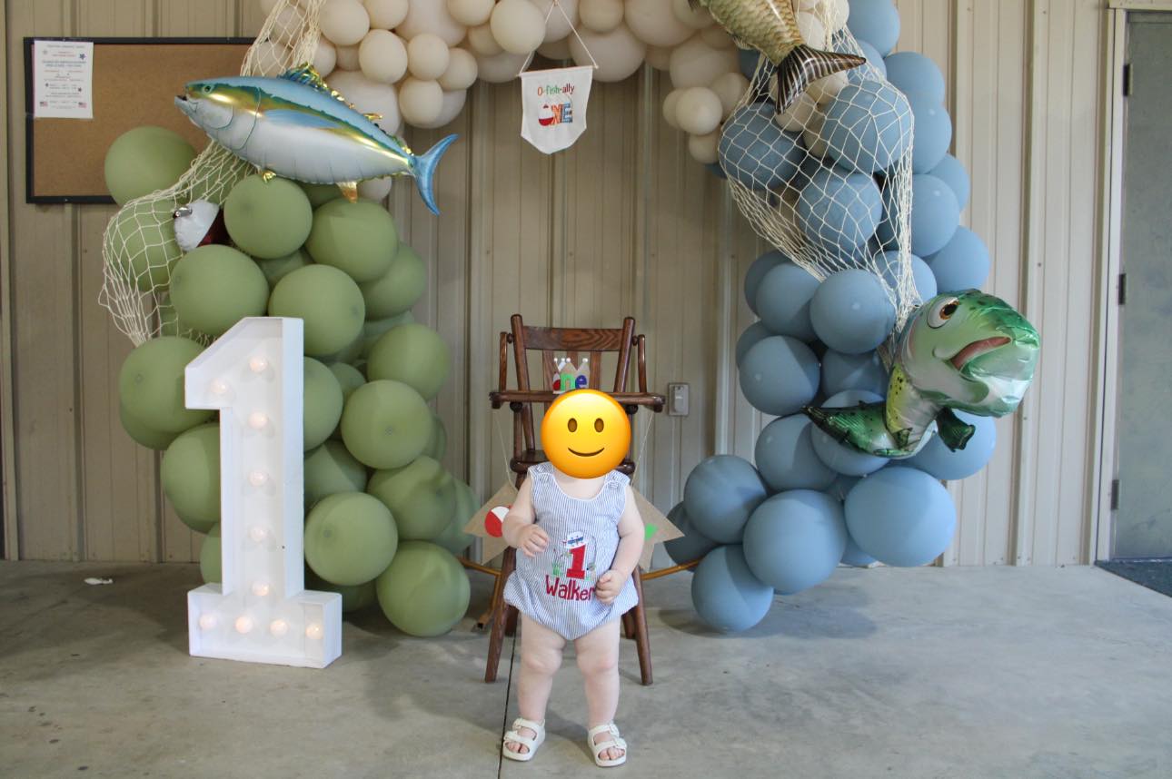 First birthday backdrop with balloon arch, fishing nets, fish balloons, and a number one marquee light.