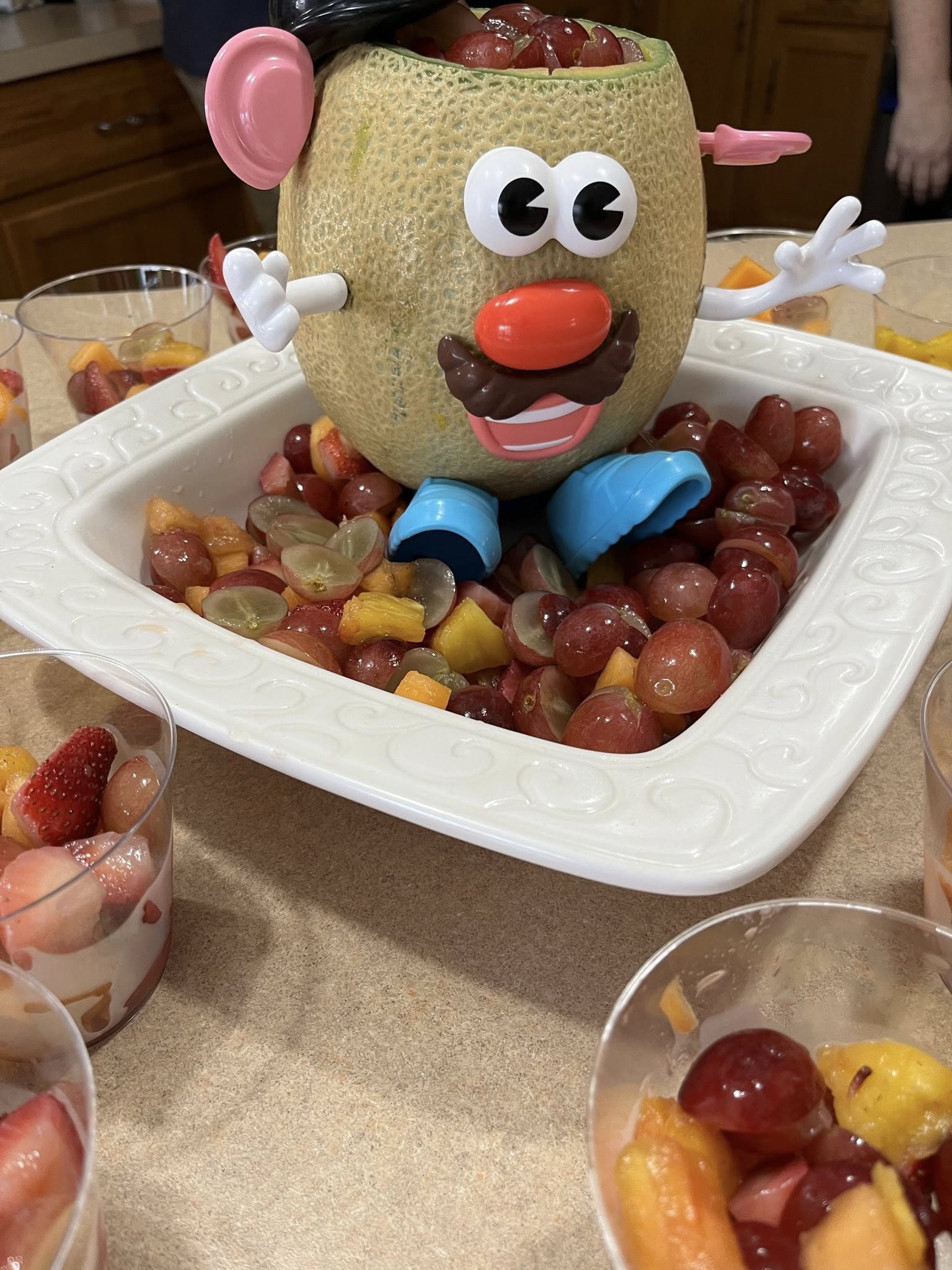 Cantaloupe carved into a Mr. Potato Head shape sitting in a bowl of fresh fruit salad.