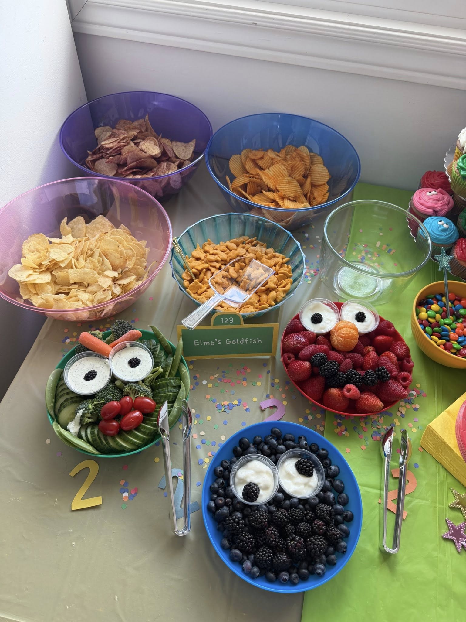Sesame Street-themed party food table with bowls of chips and snack trays shaped like Cookie Monster and Elmo.