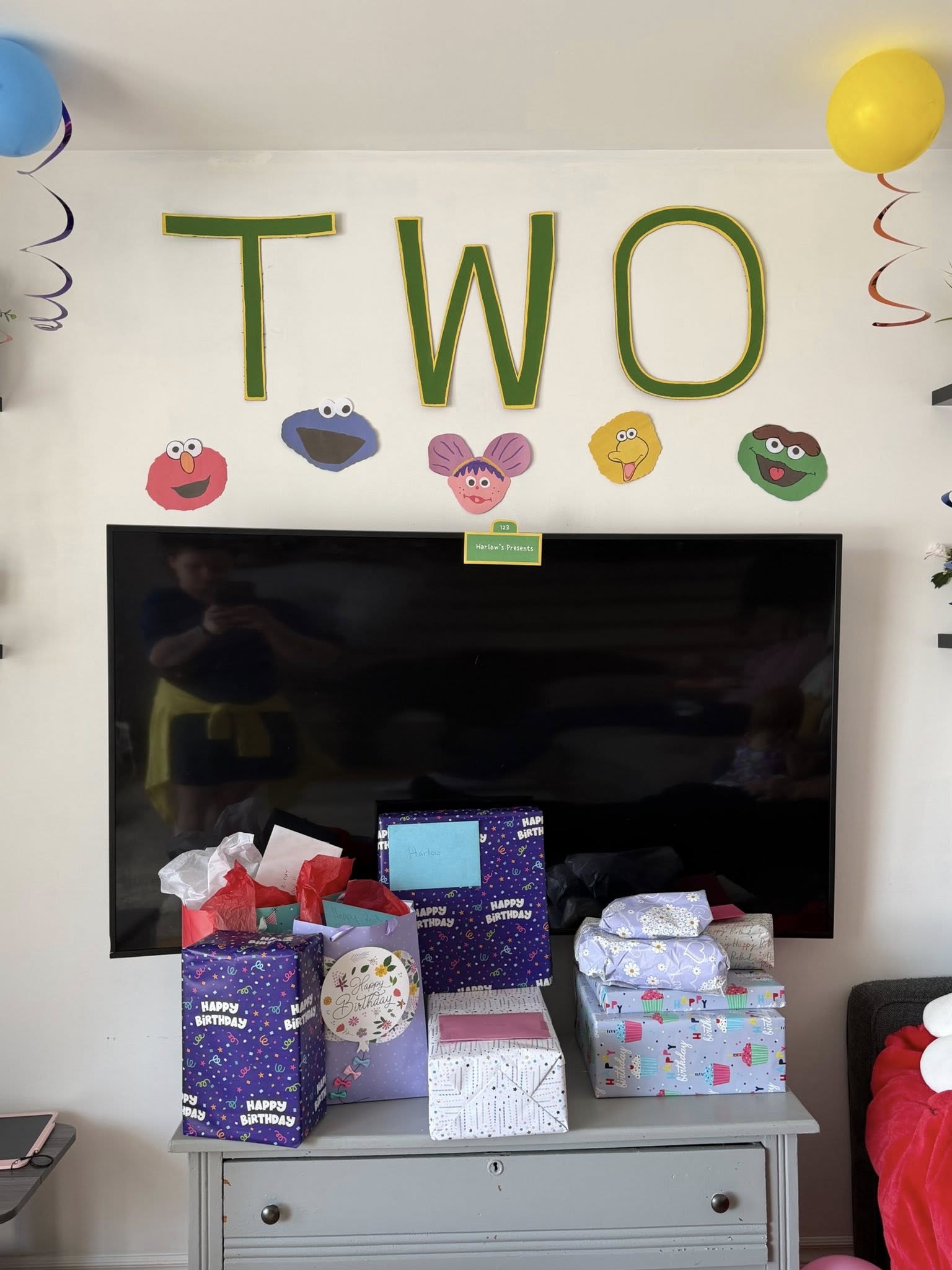Birthday gifts stacked below wall decorations spelling TWO with Sesame Street character cutouts.