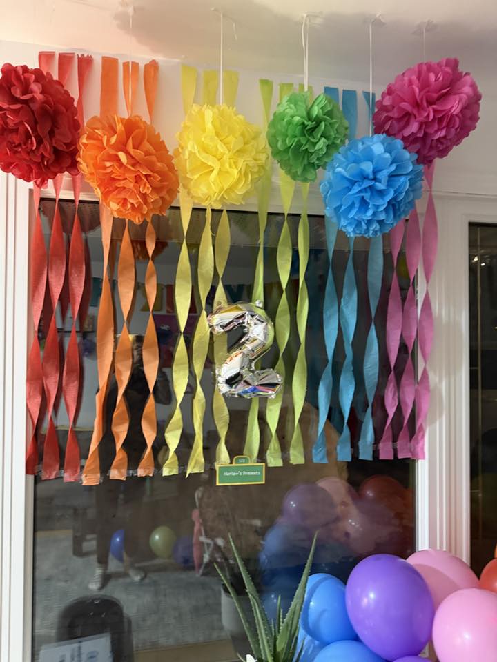 Colorful rainbow paper pom-poms and crepe streamers hanging in a window with a silver number 2 balloon decoration.