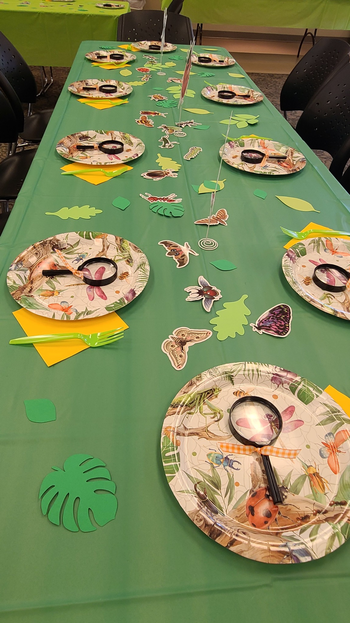 Table set for a bug-themed party with insect plates, green tablecloth, paper leaves, and toy magnifying glasses.
