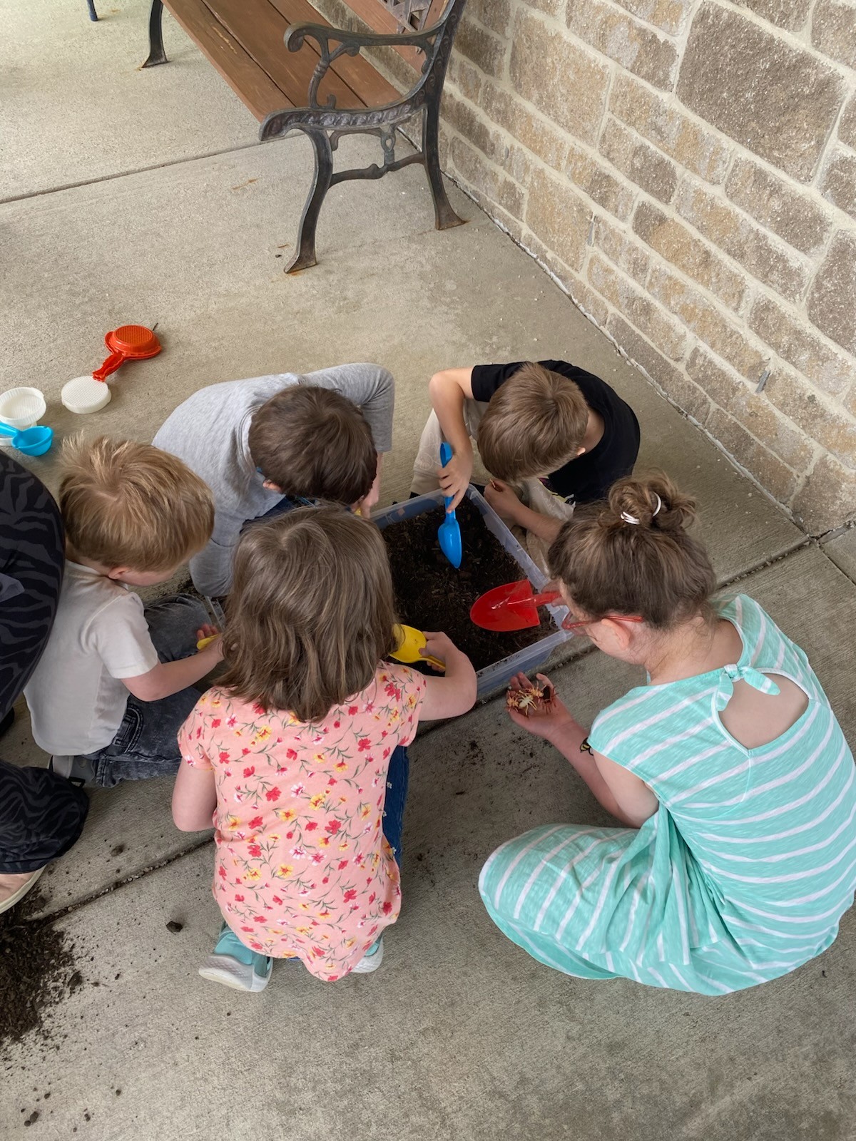 Four children sitting outside on a patio playing with toy shovels in a bin of dirt.