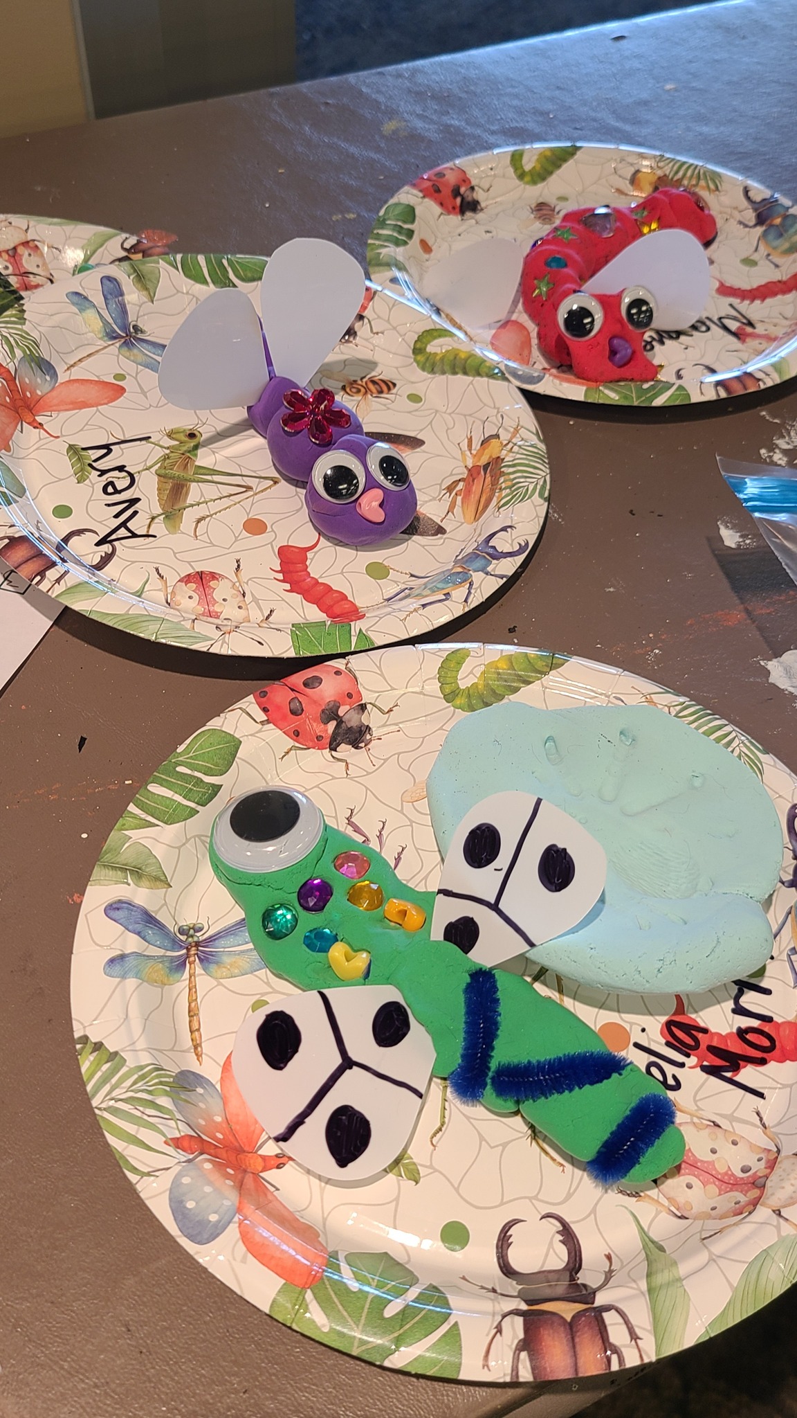 Colorful homemade insect crafts made from clay, googly eyes, and paper wings on decorative bug-themed plates.