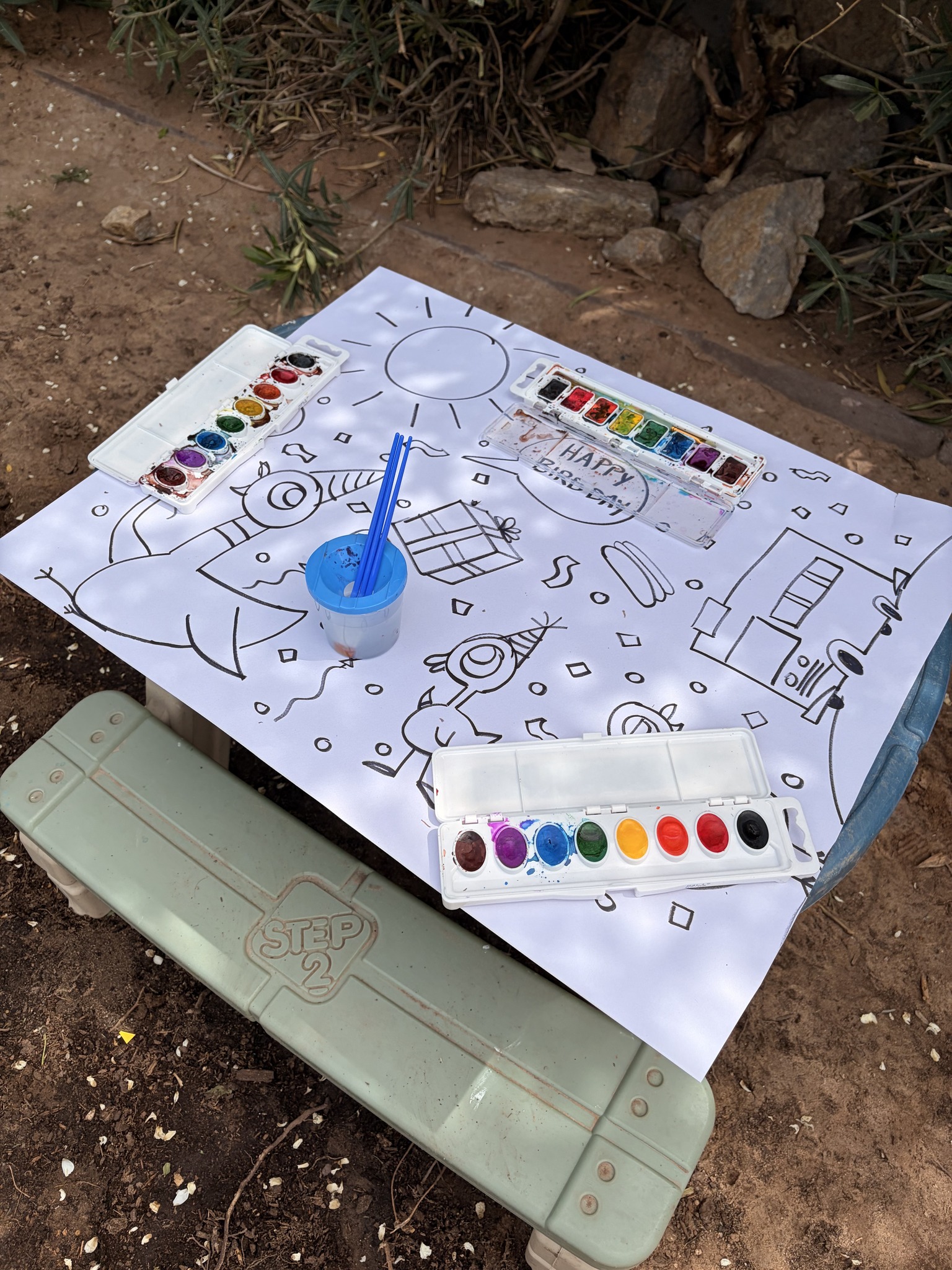 Step2 activity table with birthday-themed coloring page, watercolors, and brushes outdoors.