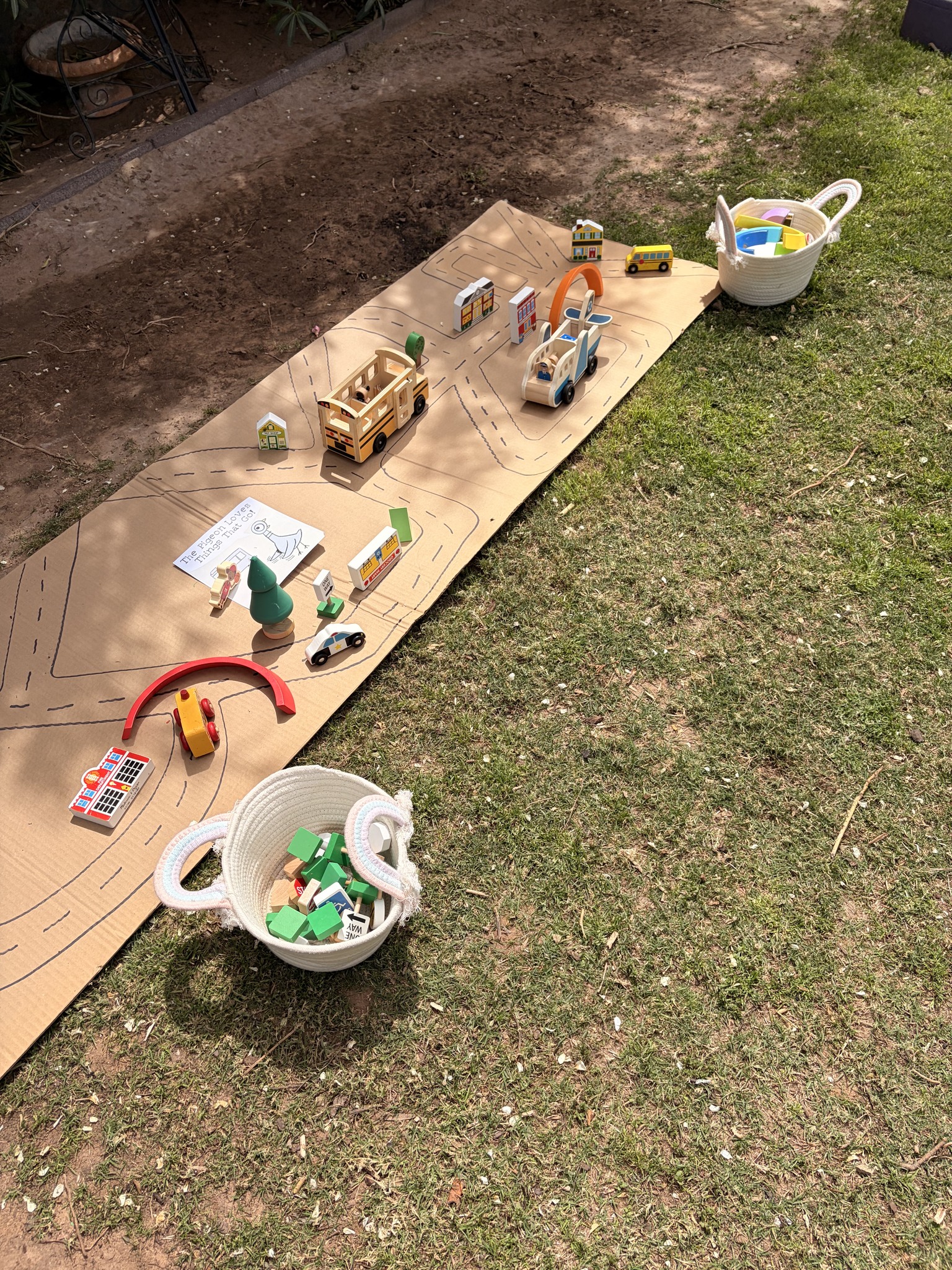 Outdoor DIY cardboard road play set with wooden toy vehicles and buildings on green grass.