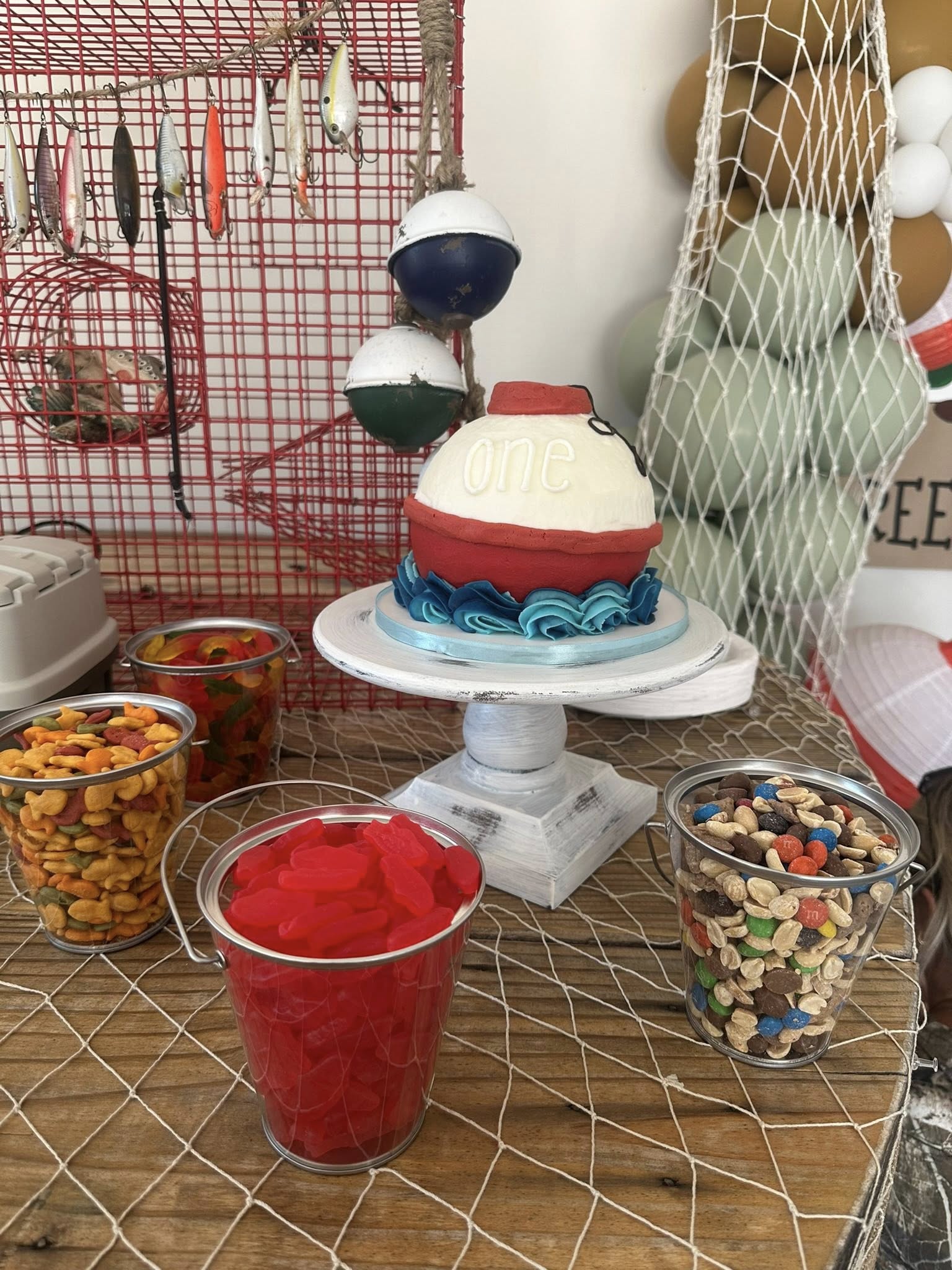 First birthday cake decorated as a fishing bobber surrounded by snack buckets on a fishing-themed party table.