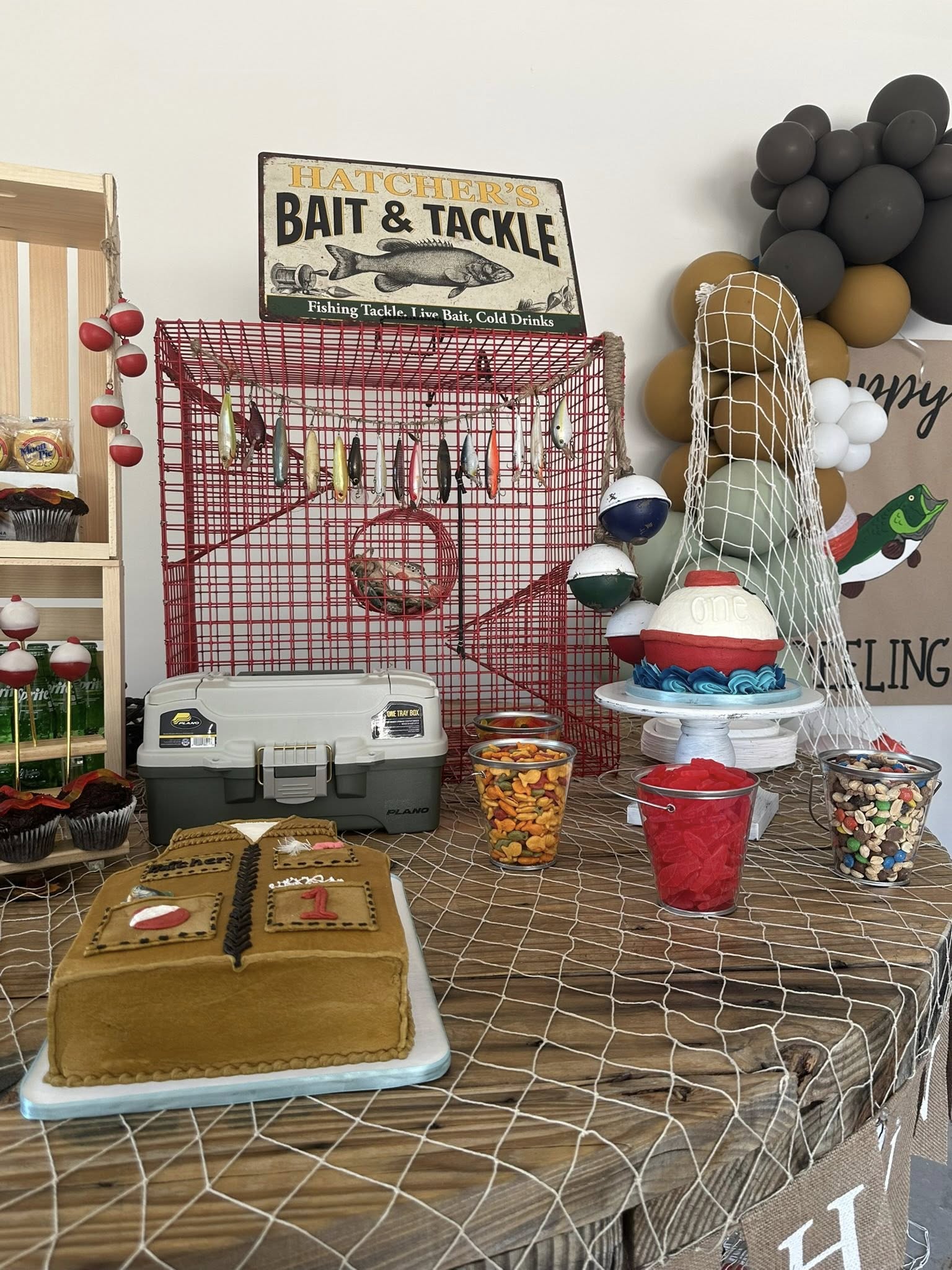 Fishing-themed first birthday party dessert table decorations with tackle box, fishing vest cake, and bobber cake.