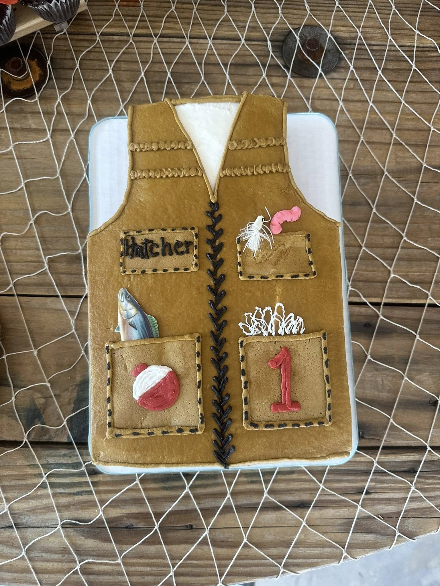 First birthday cake decorated as a tan fishing vest with pockets, a red bobber, a fish, and the name Hatcher.