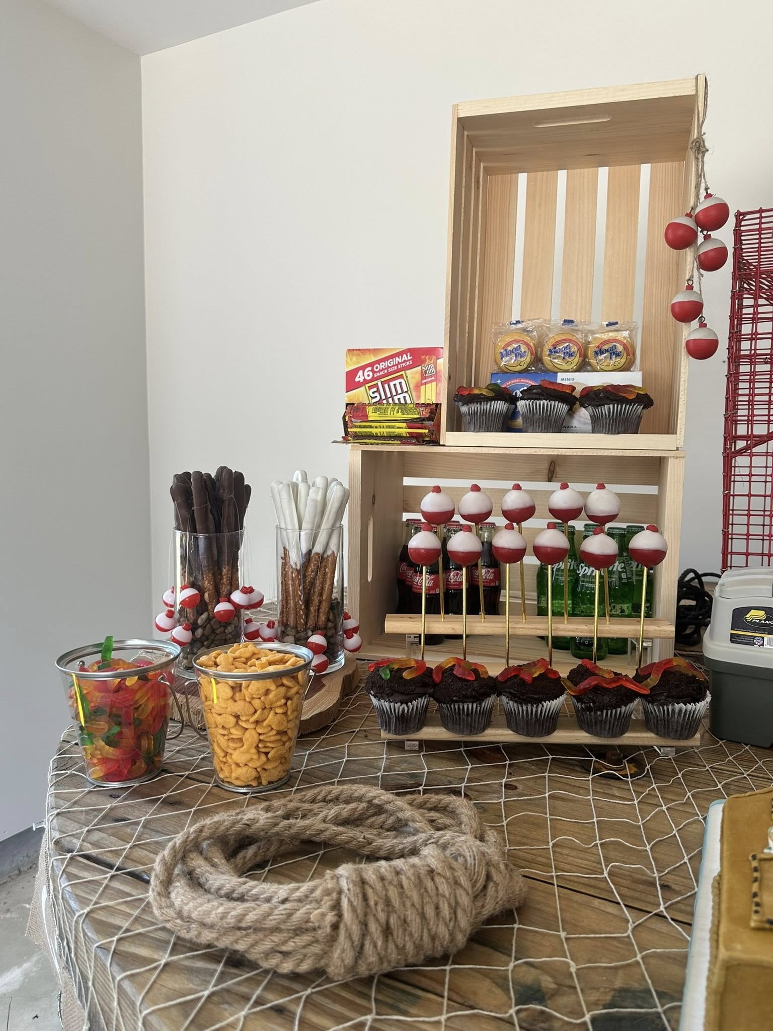 Fishing themed snack table featuring chocolate-dipped pretzels, gummy worms, goldfish crackers, and cupcakes with bobber-shaped cake toppers.