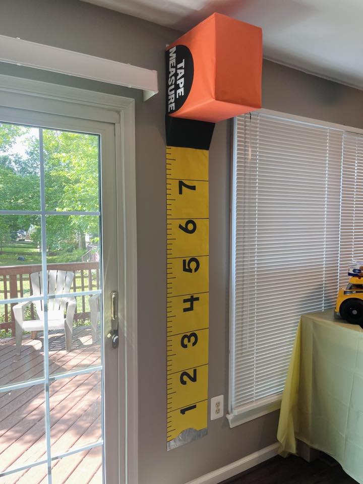 Large yellow wall-mounted tape measure prop for construction-themed party, positioned near a window.