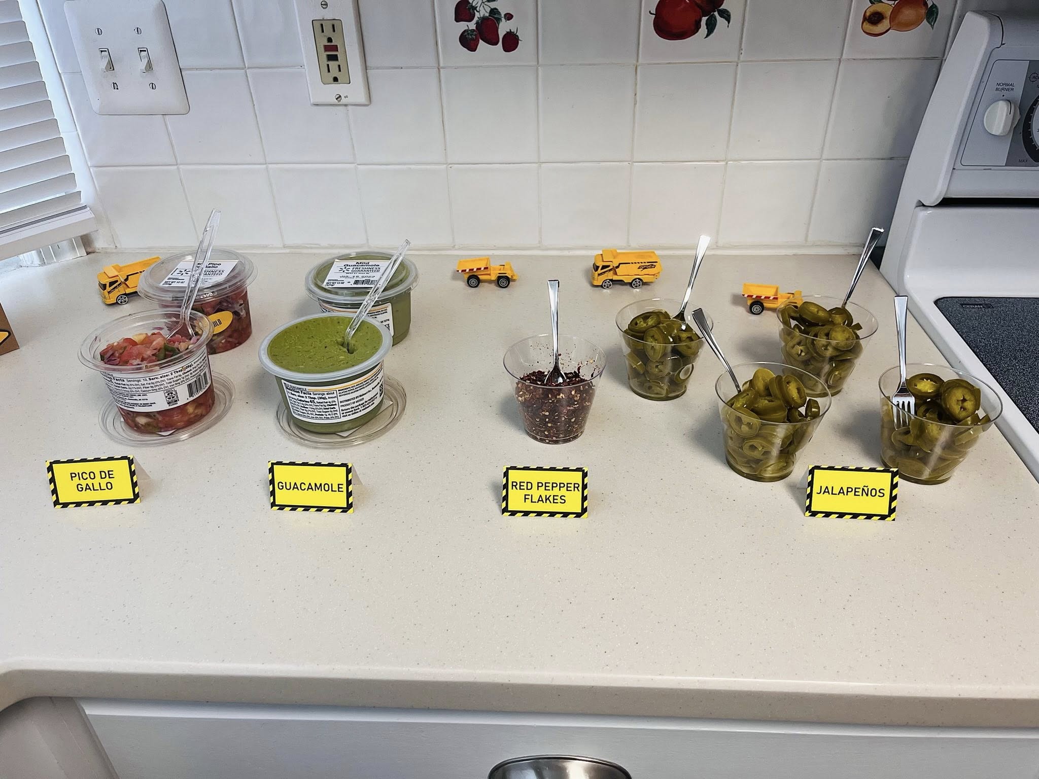 Table spread of taco bar condiments including pico de gallo, guacamole, red pepper flakes, and jalapeños.