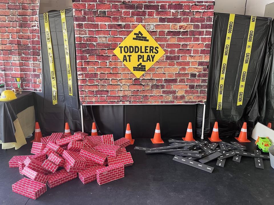 Construction-themed party backdrop with a Toddlers at Play sign, caution tape, toy bricks, and orange traffic cones.