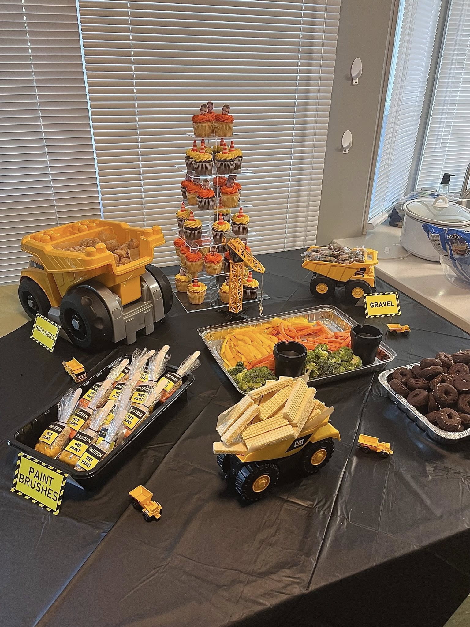 Construction-themed party table featuring dump trucks serving food labeled as boulders, gravel, and paint brushes.
