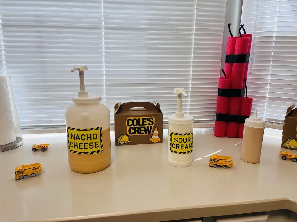 Construction-themed party table set with condiment dispensers labeled Nacho Cheese and Sour Cream with toy dump trucks.