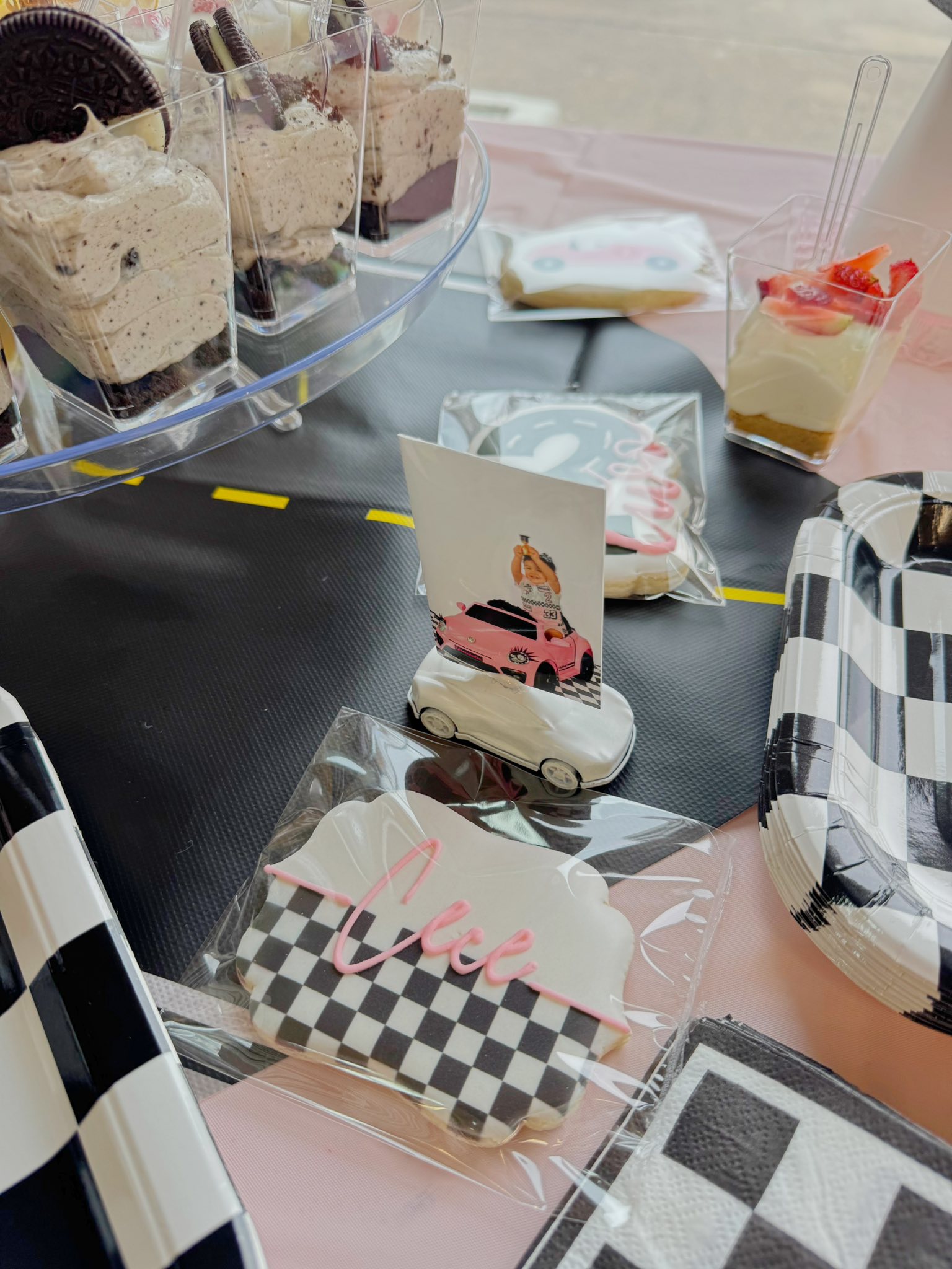 Race car-themed party treats with checkered pattern cookies, photo topper on a toy car, and dessert cups.