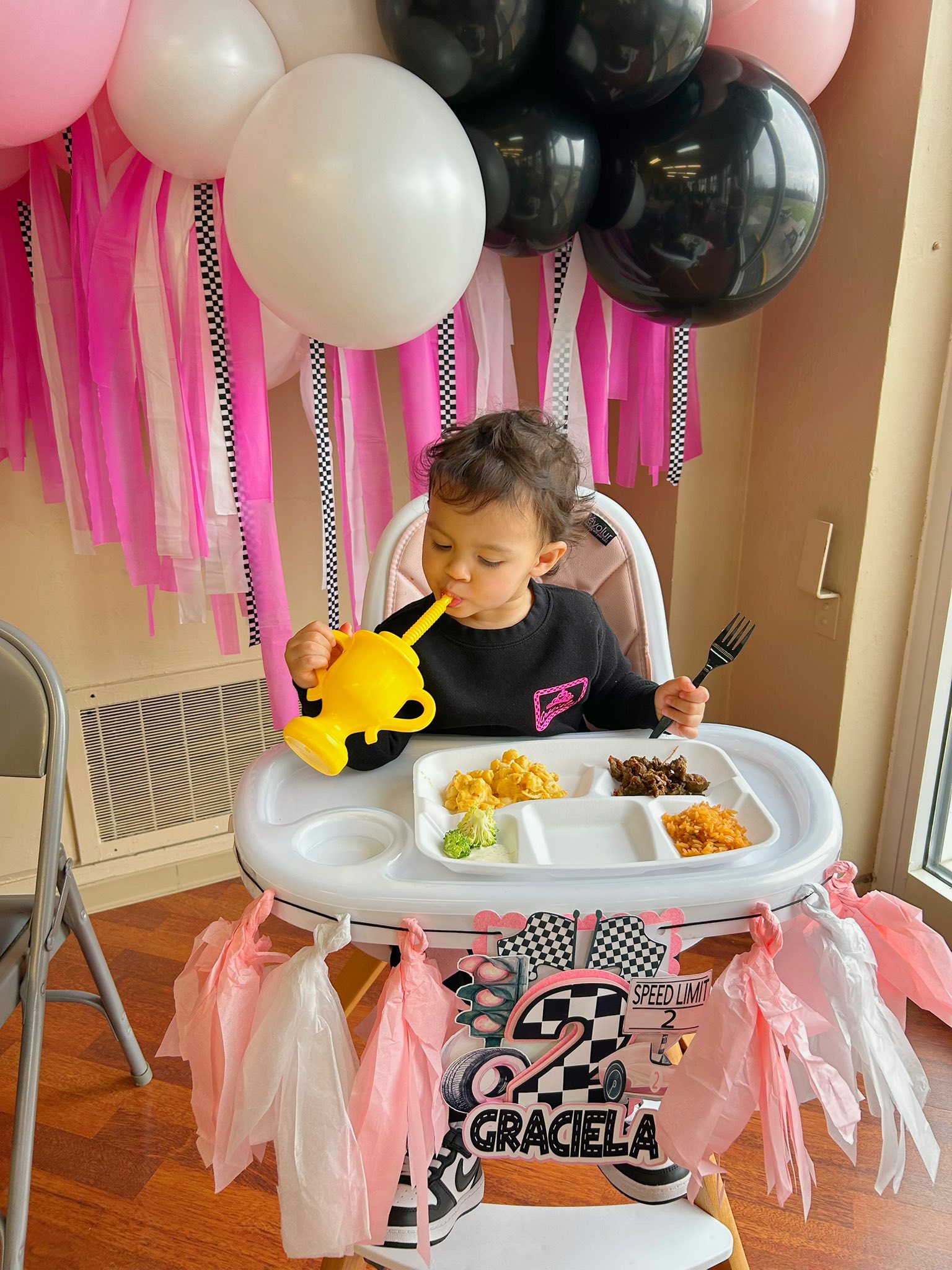 Toddler in a black shirt sitting in a high chair at a race car themed birthday party, drinking from a trophy cup.