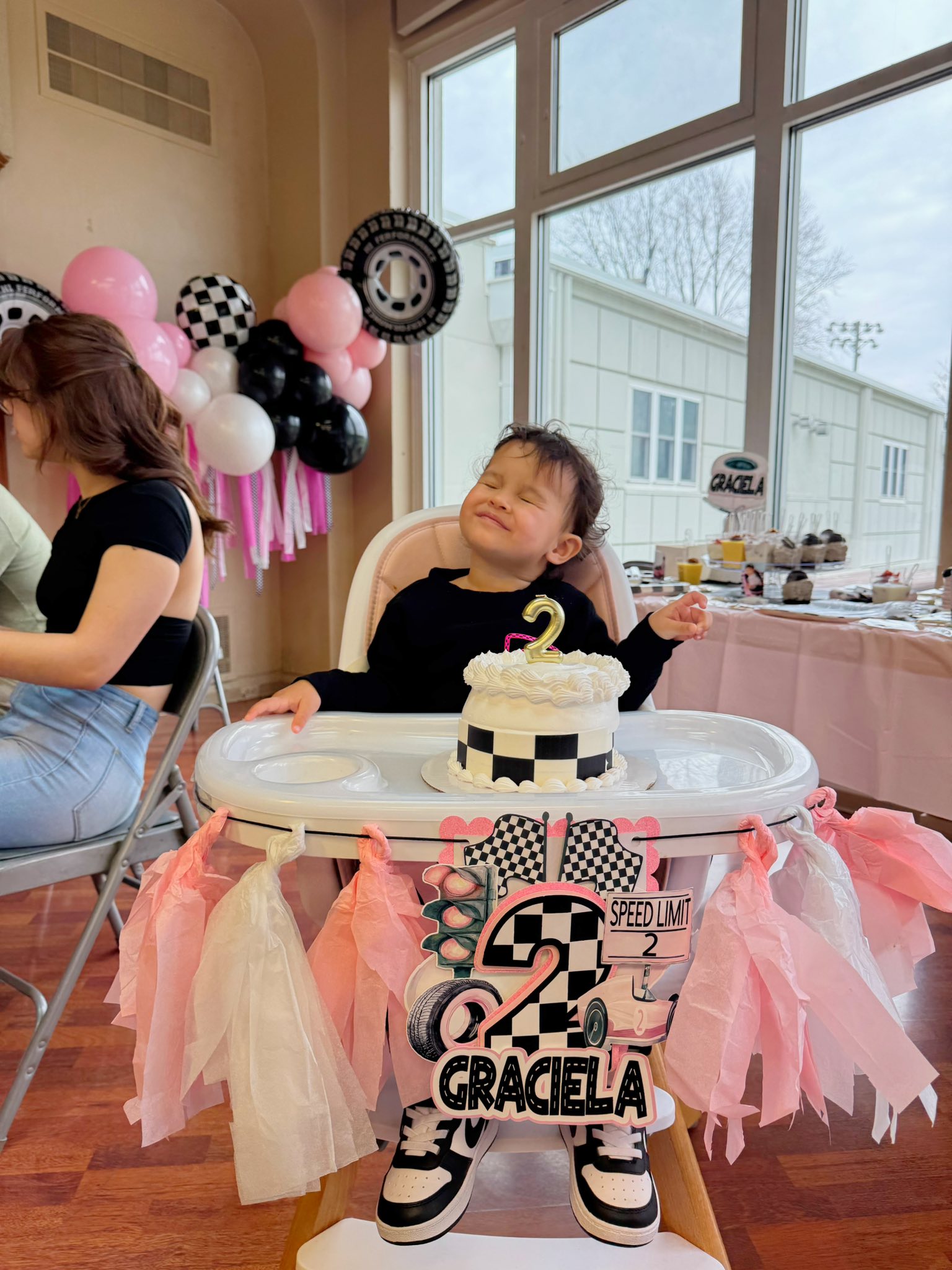 Toddler in a high chair at a racing-themed second birthday party with a themed cake and personalized decorations.