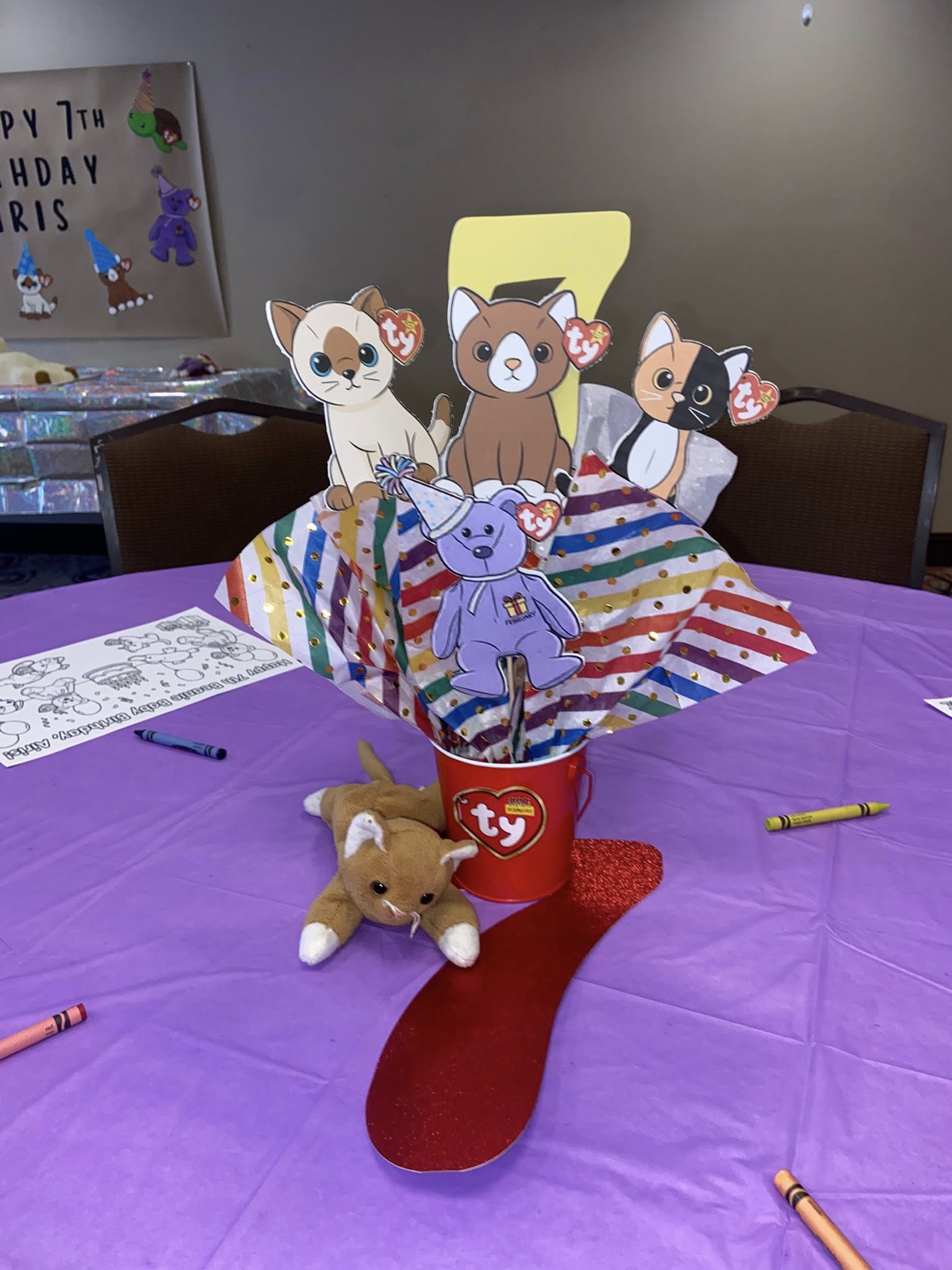 Beanie Baby themed birthday centerpiece with a plush cat, Ty bucket, and character cutouts on a purple tablecloth.