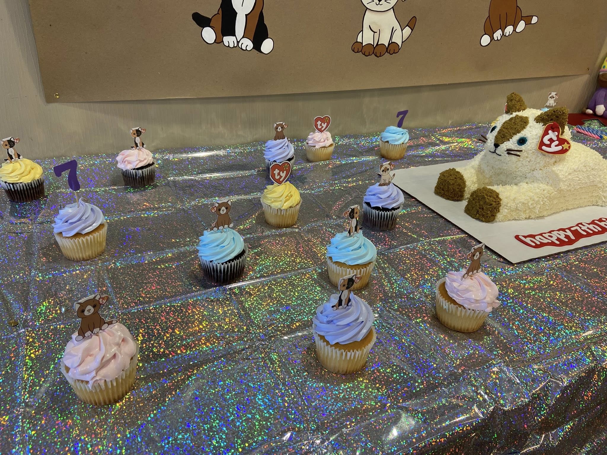 Cat-themed birthday cupcakes with Ty plushie toppers and a cat-shaped cake on a holographic party table