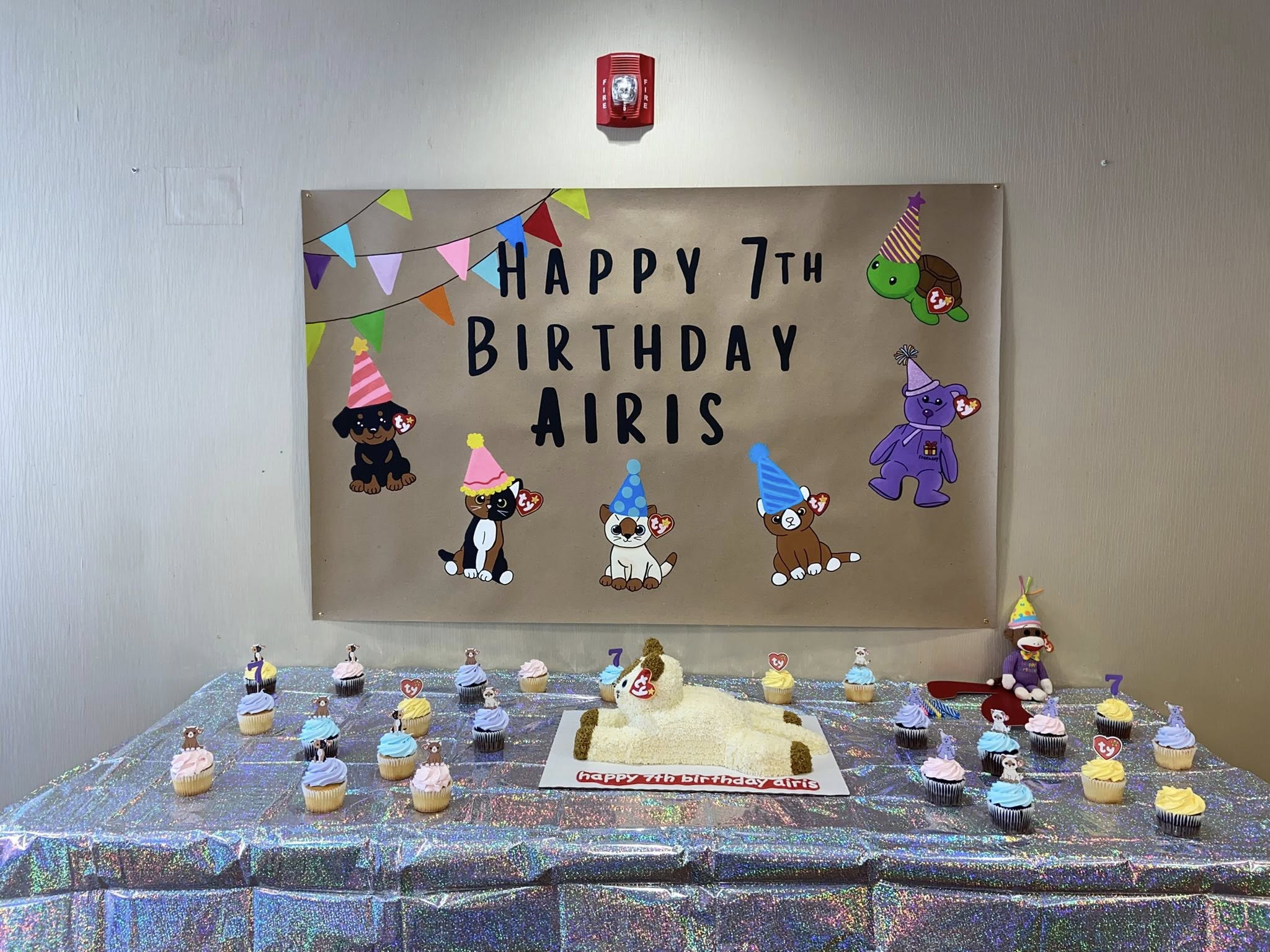 Beanie Baby themed birthday party setup with a wall banner and table decorated with cupcakes and cat-shaped cake.