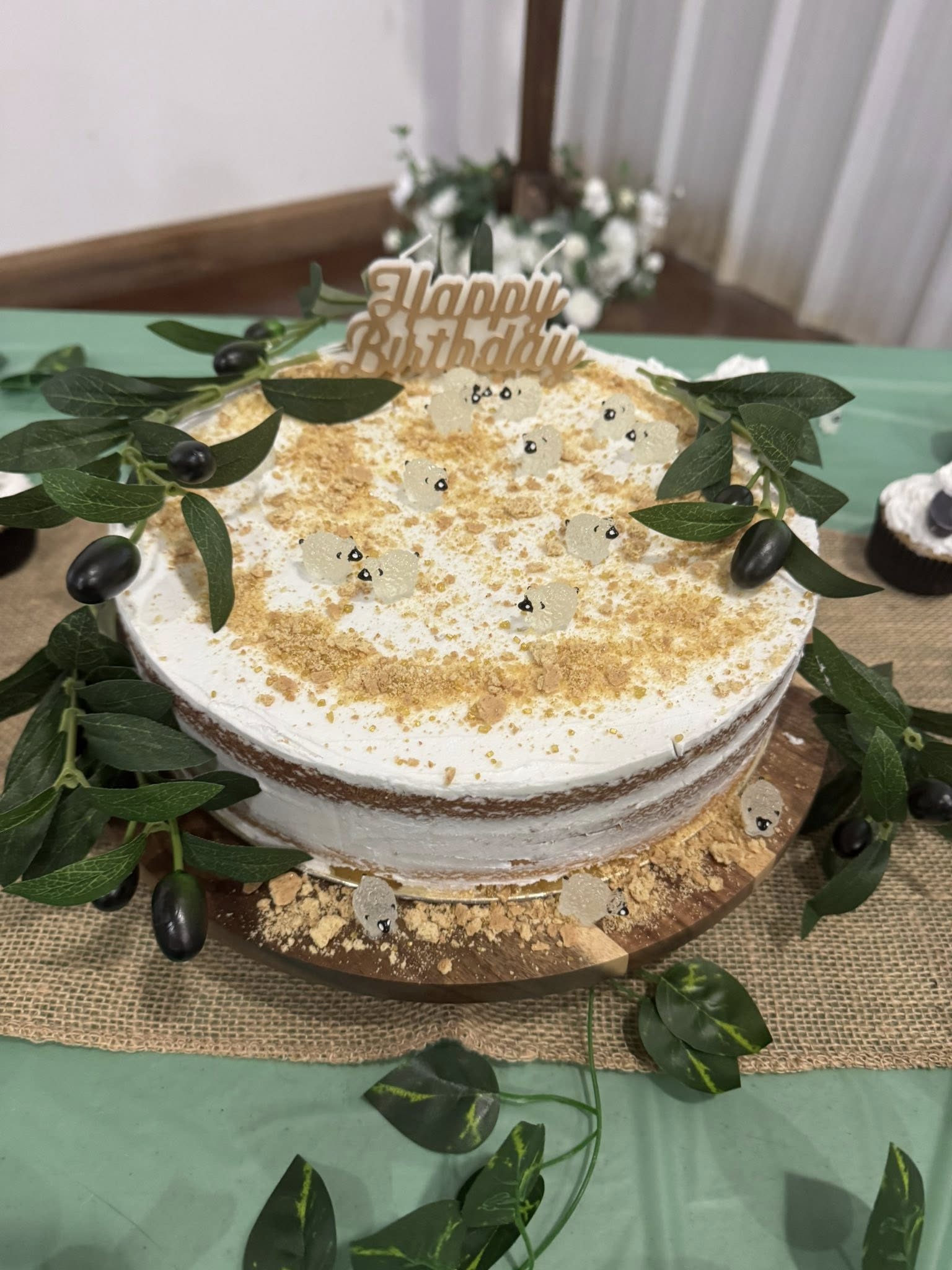 Naked sponge cake with white frosting, crumble topping, and sheep-shaped fondant decorations, adorned with a Happy Birthday topper and olive branch accents on a wooden serving board.