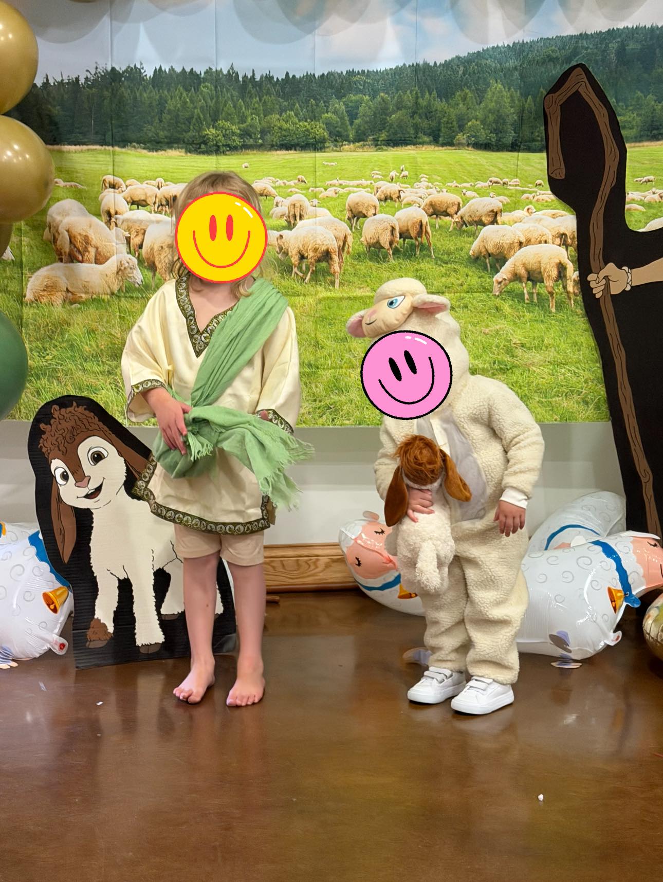 Two children dressed in shepherd and sheep costumes in front of a rustic landscape backdrop featuring a flock of sheep.