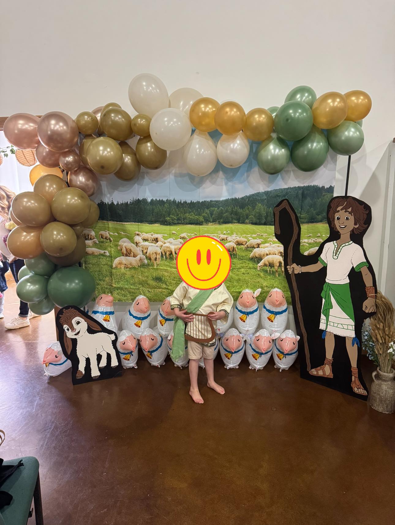 Child dressed as a shepherd with a harp at a Bible-themed party with sheep balloons and character cutouts.
