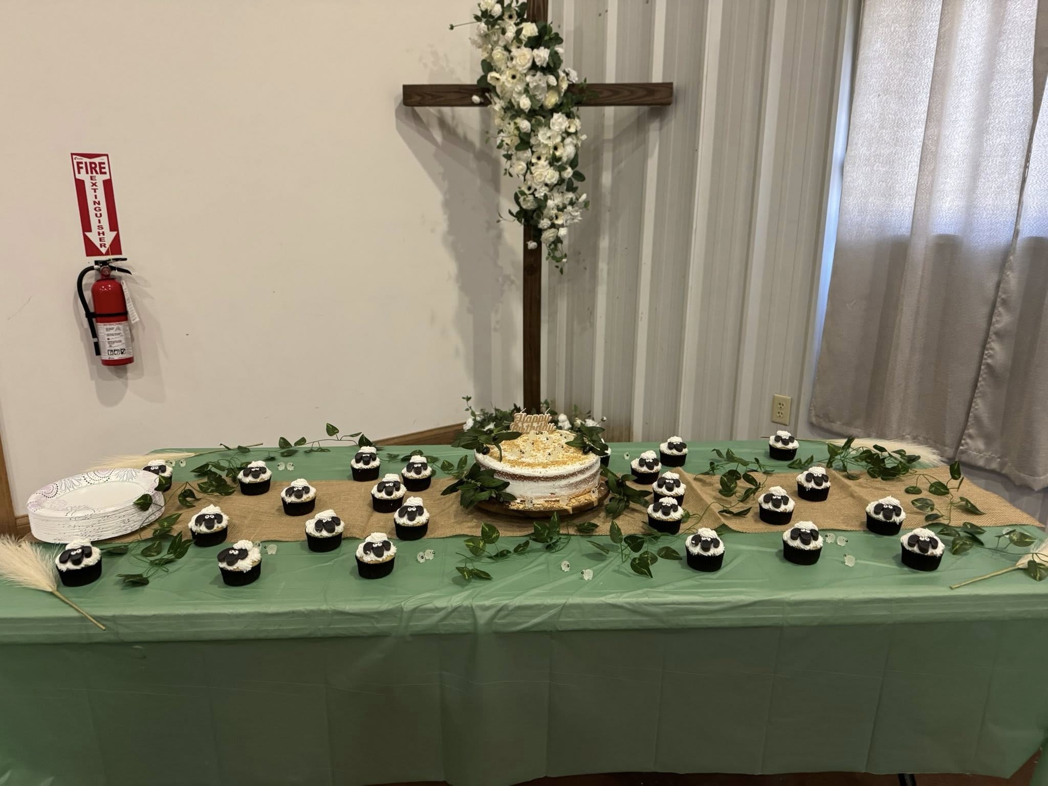 Birthday table featuring a cake and sheep-themed cupcakes decorated with greenery against a wooden cross background.
