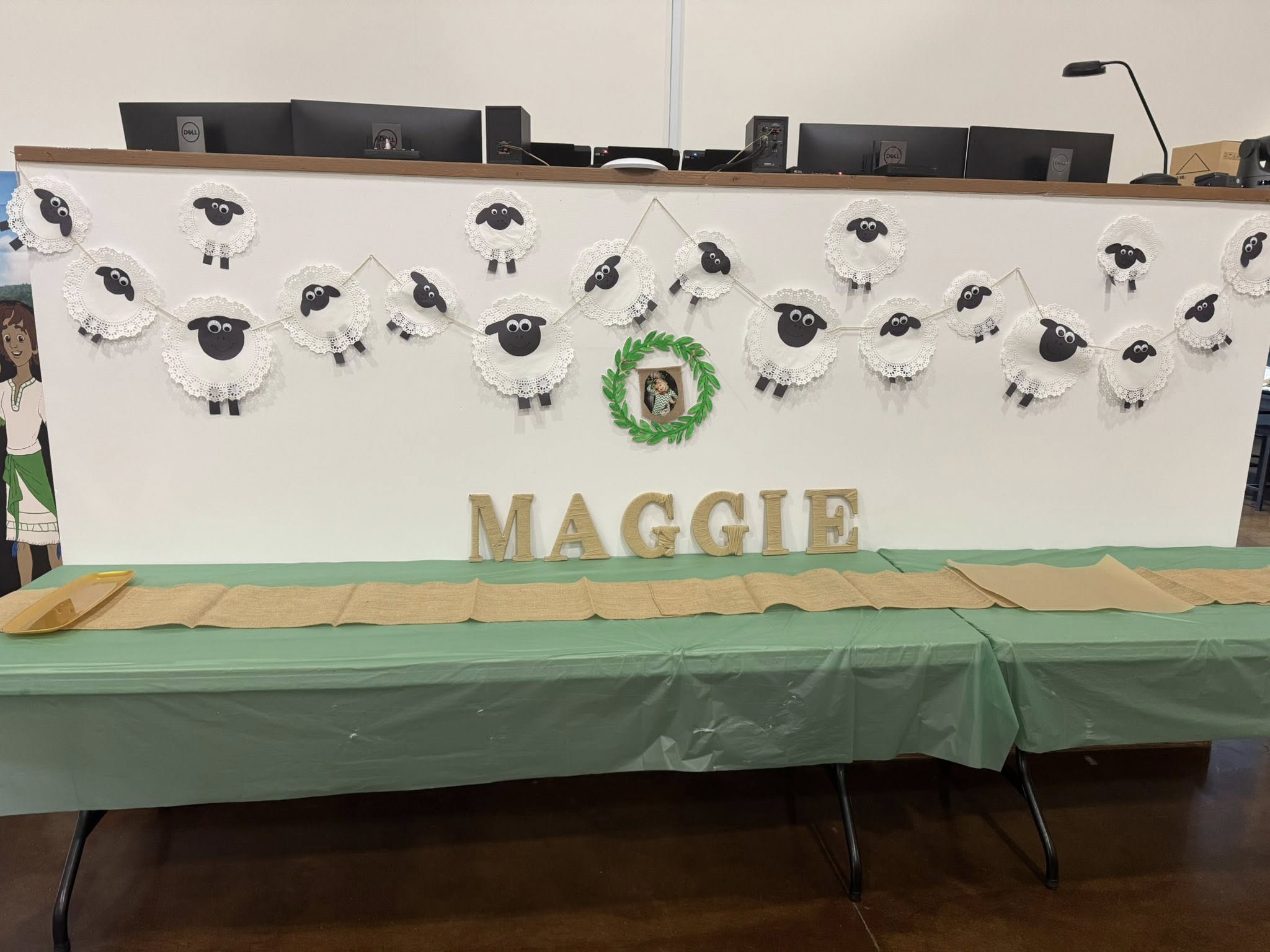 Sheep themed party decorations with a garland of paper doily sheep and wooden MAGGIE lettering above a green table