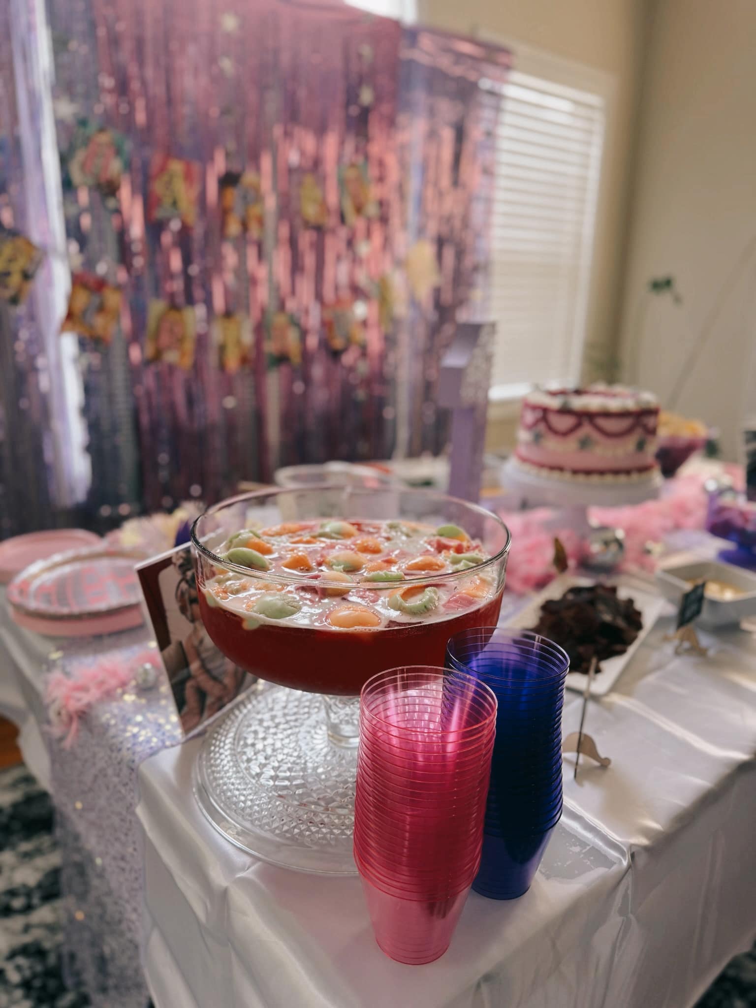Glass punch bowl filled with pink punch and fruit scoops, with stacks of pink and blue plastic cups nearby.