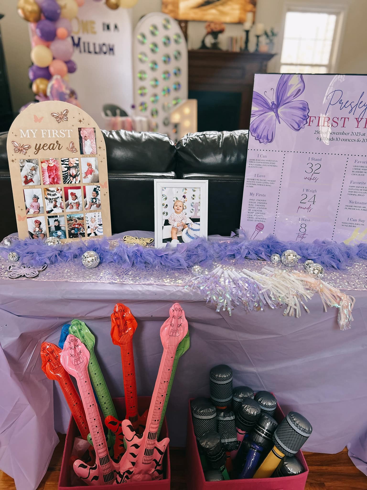 First birthday party table with a milestone photo collage, baby portrait, and information sign, plus inflatable guitars and microphones for party favors.
